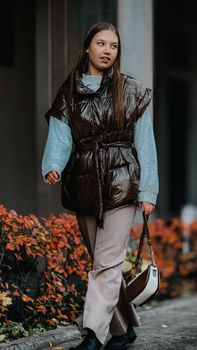 Stylish young woman in brown vest and grey sweater walking with handbag outdoors.