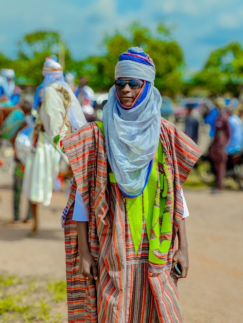 Free Man in traditional African attire, wearing sunglasses and a turban at a lively outdoor event. Stock Photo