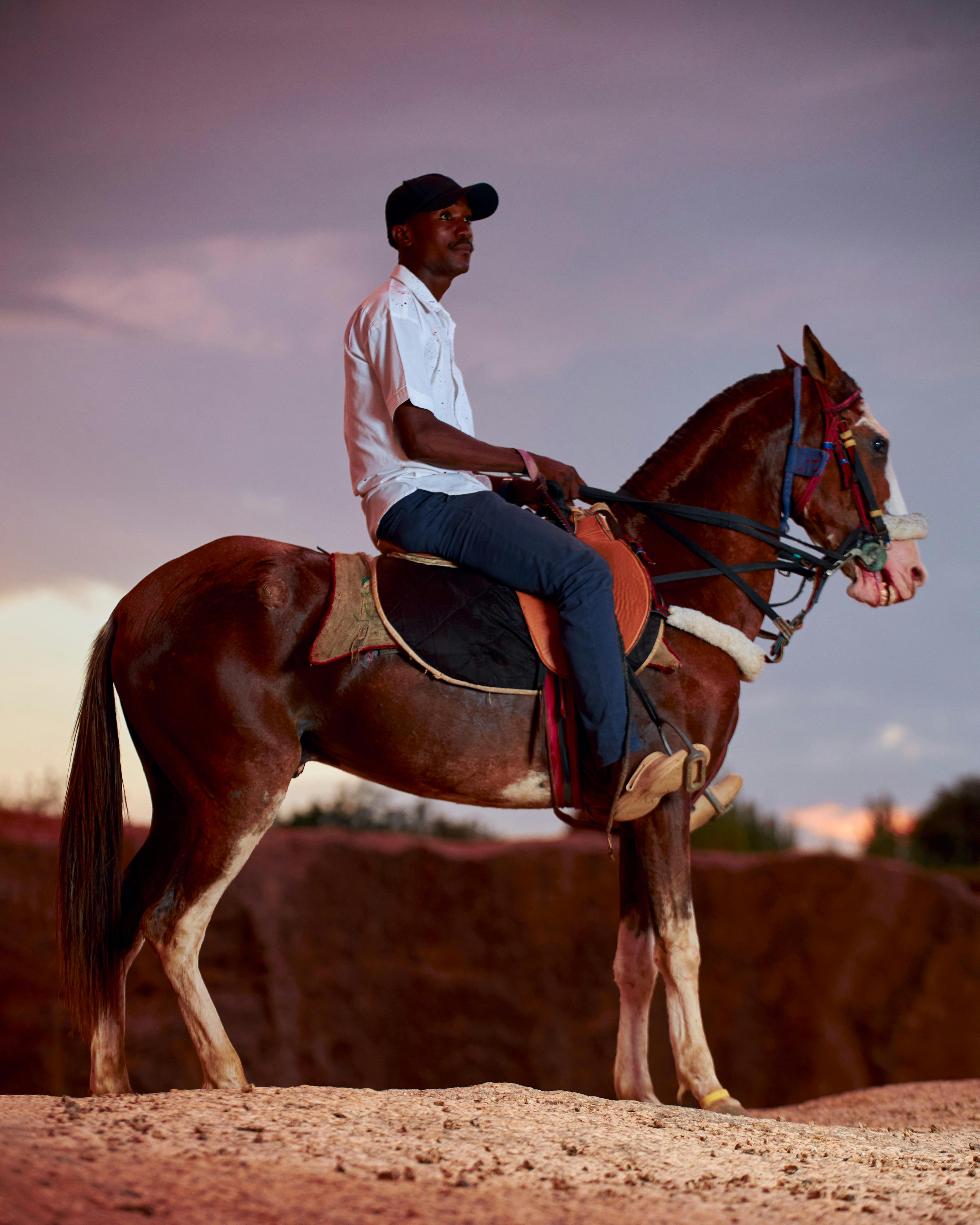 A man in white shirt and cap riding a horse at sunset in a desert landscape.