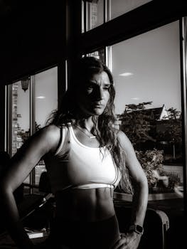 Confident woman in a gym setting, highlighted by sunlight and shadows, showcasing strength.
