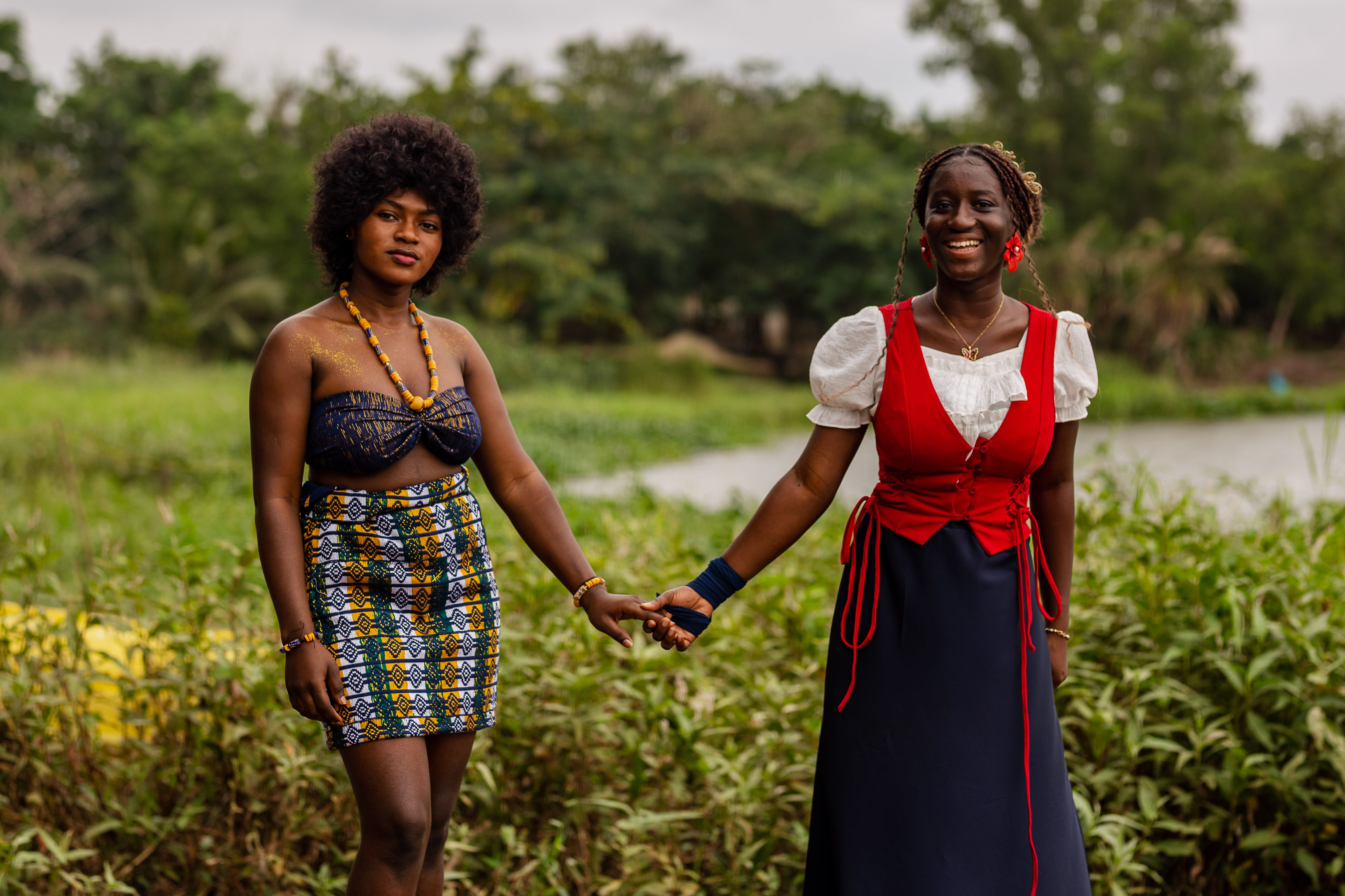 Two African women in traditional attire joyfully holding hands in a lush garden setting.