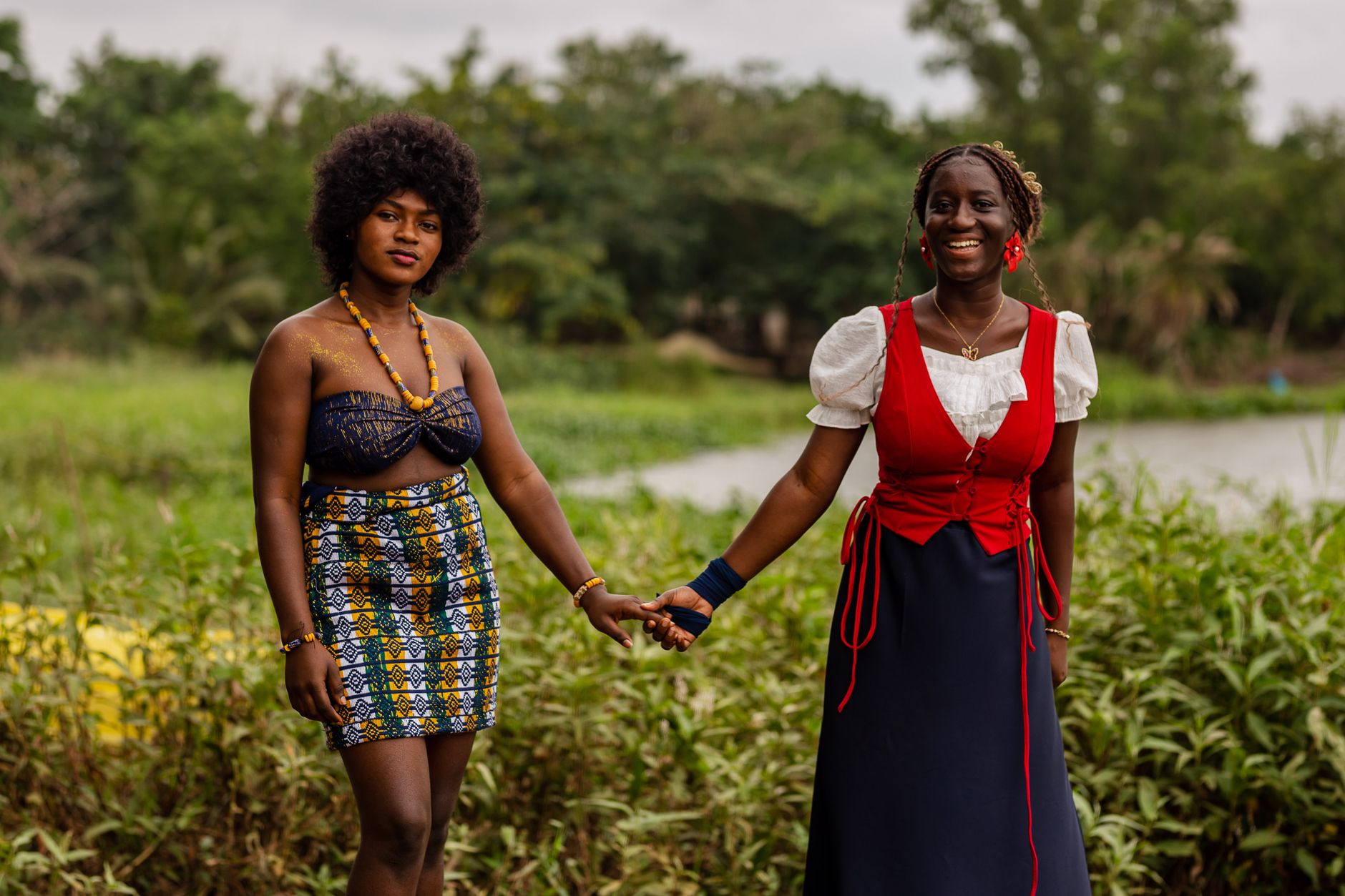 Two African women in traditional attire joyfully holding hands in a lush garden setting.