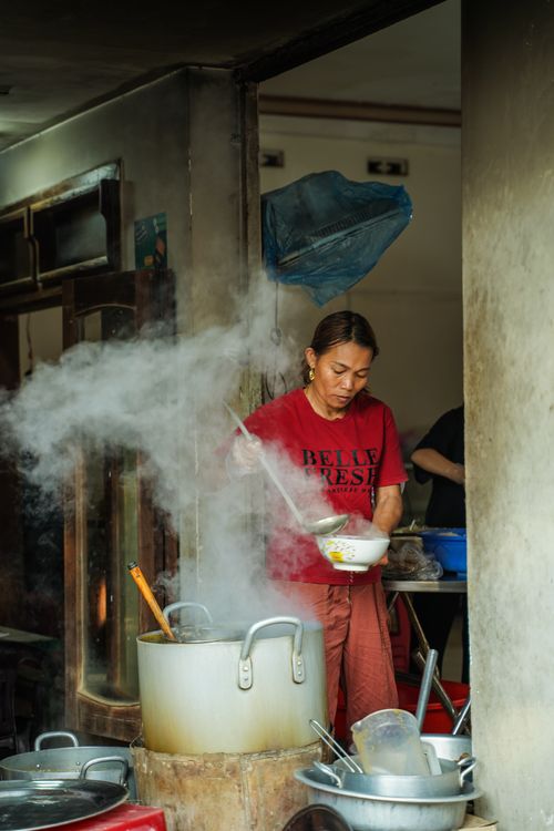 Free Female street vendor prepares steaming noodle soup in an urban market setting. Stock Photo