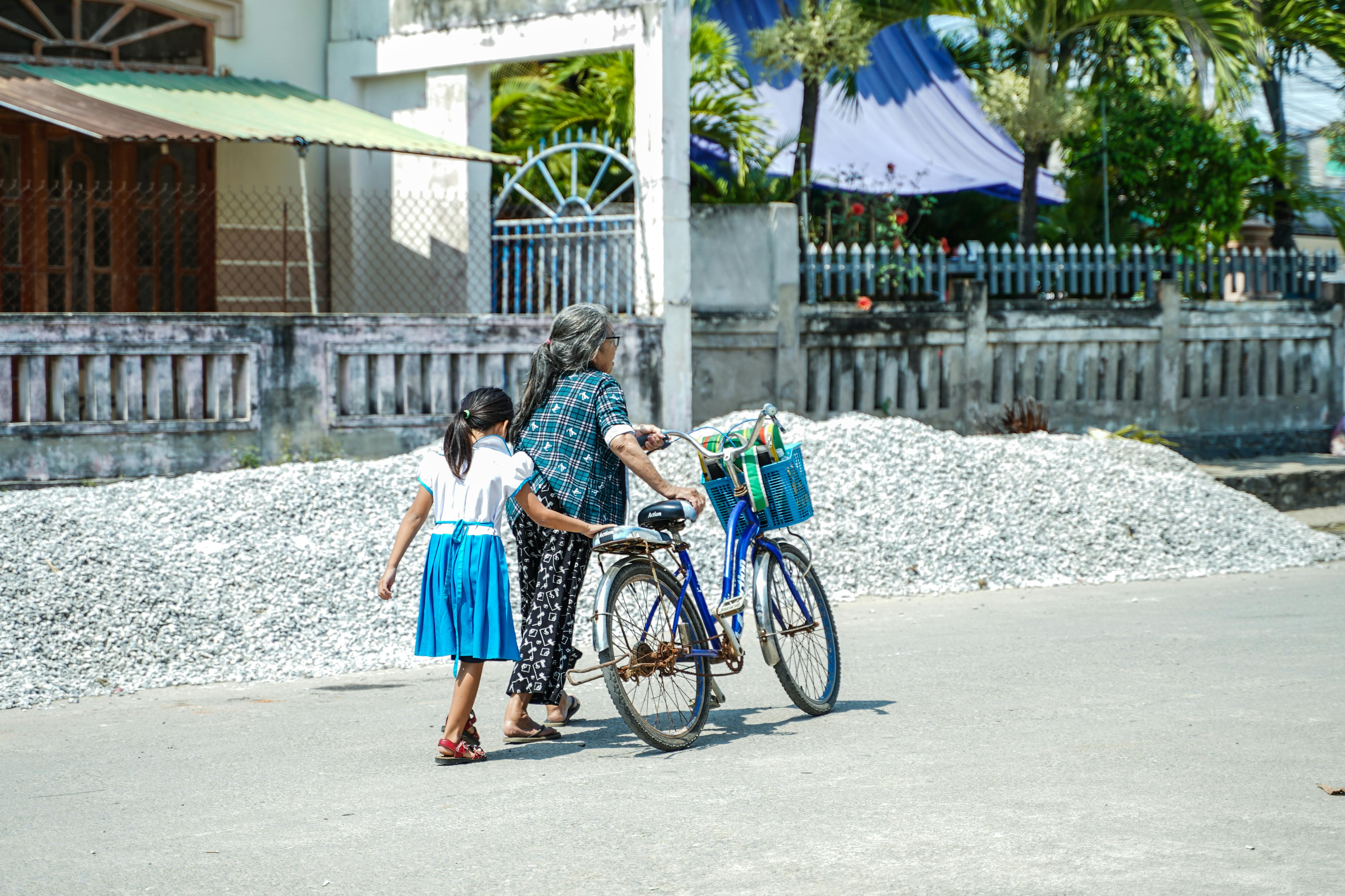 An elderly woman with a child walks along the street, guiding a bicycle on a sunny day.