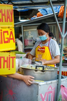 A street food vendor wearing a mask prepares a traditional dish outdoors with vibrant signage.