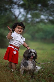 Adorable baby girl standing with a playful Shih Tzu dog in a green park, both radiating joy.