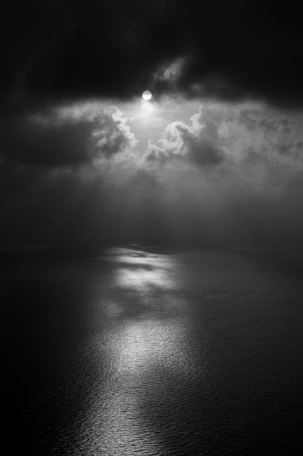 A moody black and white seascape from Corfu, Greece, featuring dramatic clouds and sunlight dancing on the water.