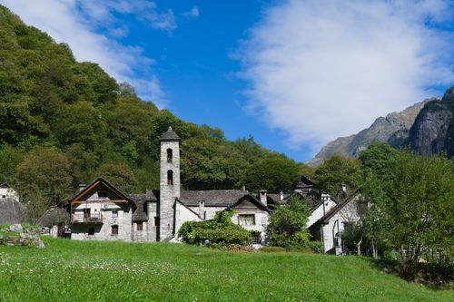 Free Charming Swiss village with stone houses and scenic Alps in the background, surrounded by lush greenery and clear blue sky. Stock Photo