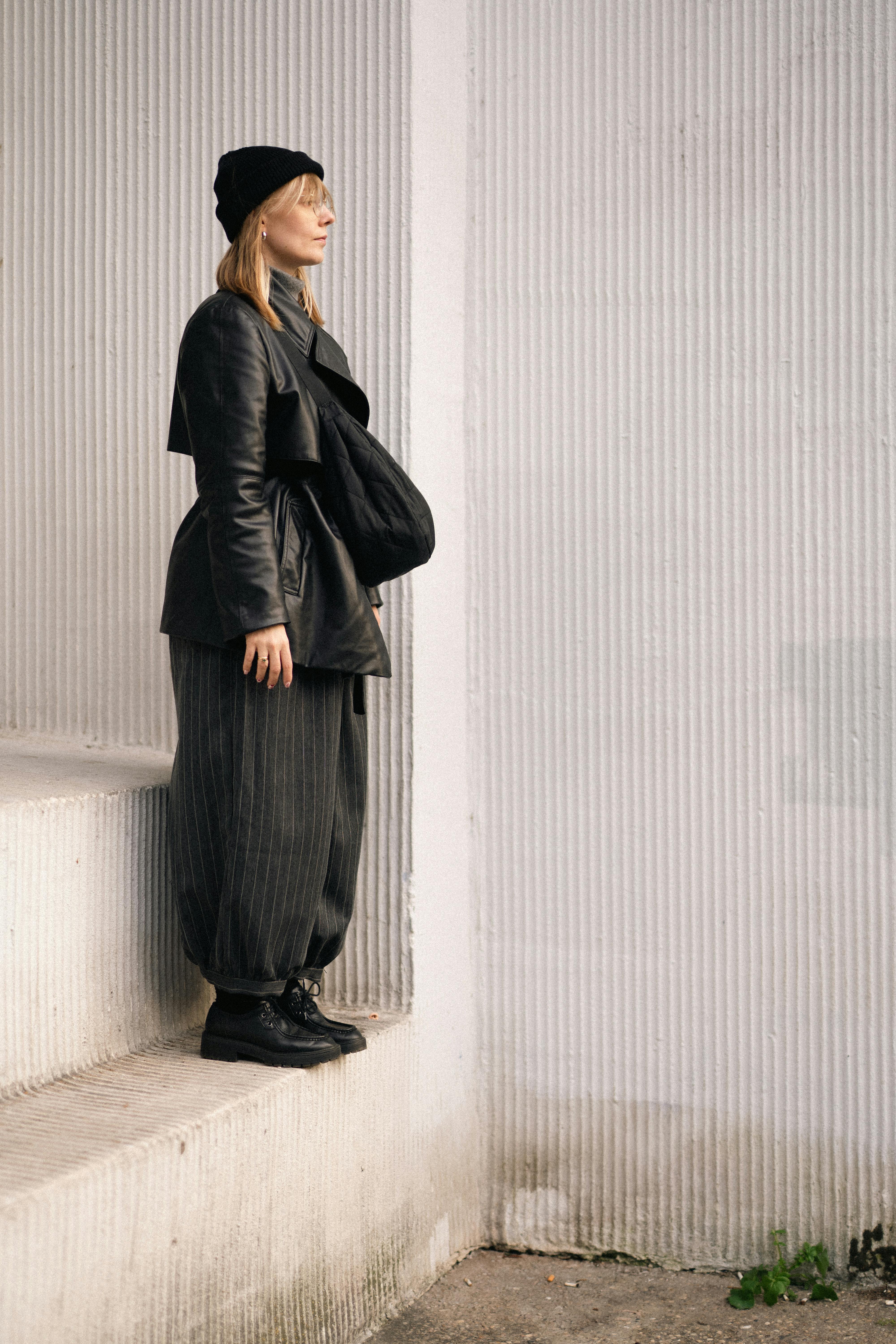 Stylish woman in contemporary clothing stands against a minimalist wall outdoors.