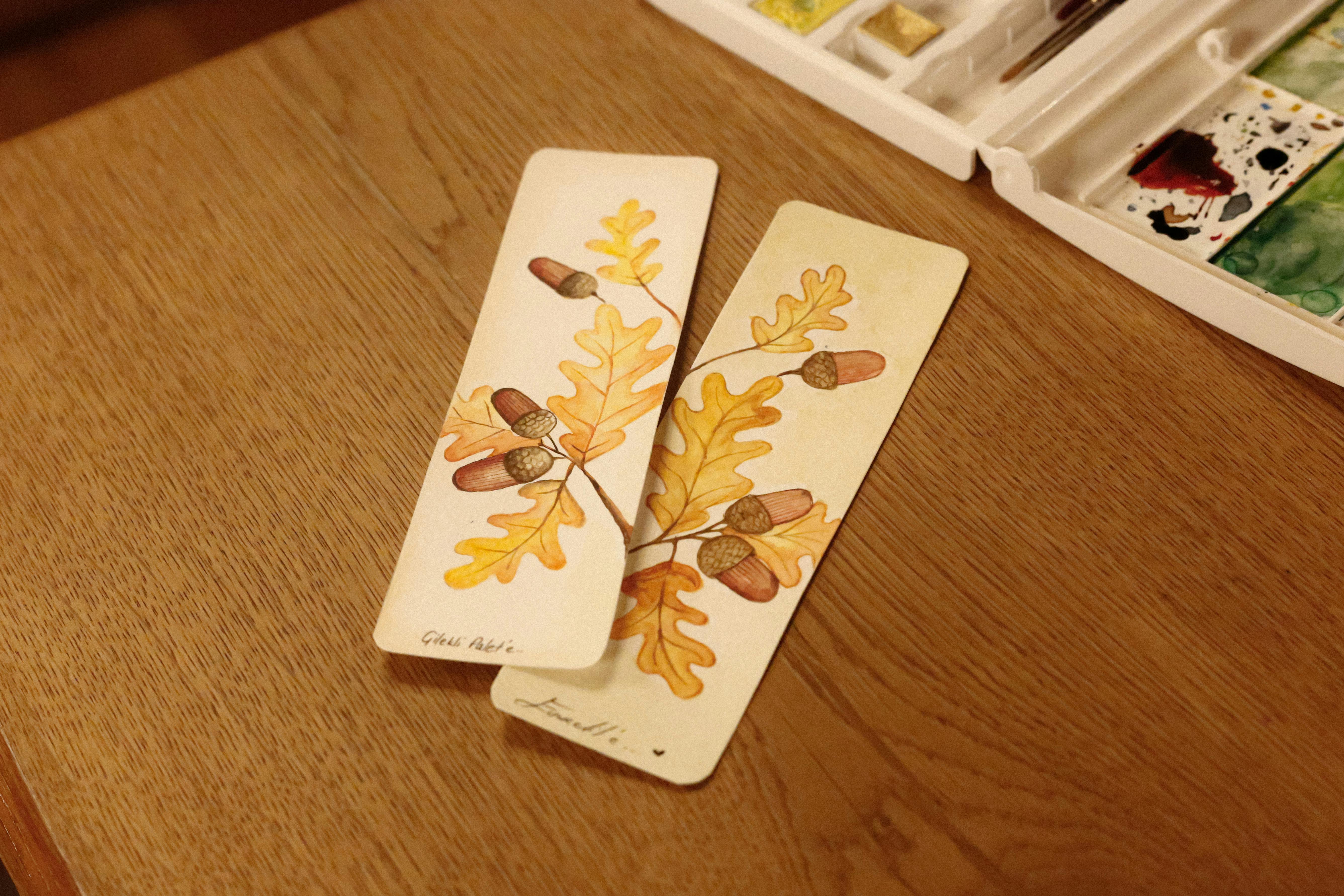 Artistic bookmarks featuring watercolor oak leaves and acorns on a wooden table.