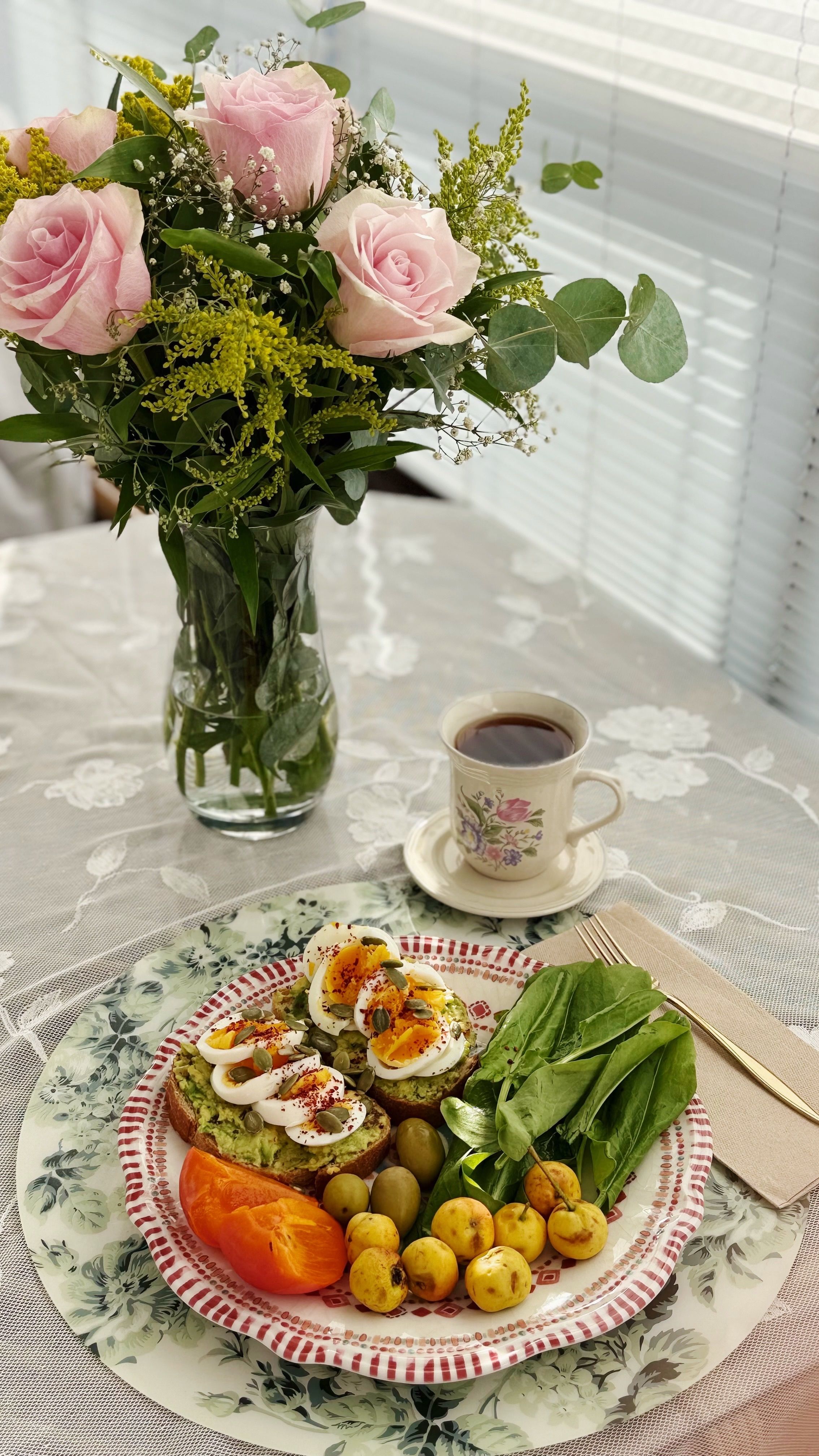 A tasteful breakfast spread with roses, coffee, avocado toast, and fruit on a lace tablecloth.