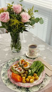 A tasteful breakfast spread with roses, coffee, avocado toast, and fruit on a lace tablecloth.