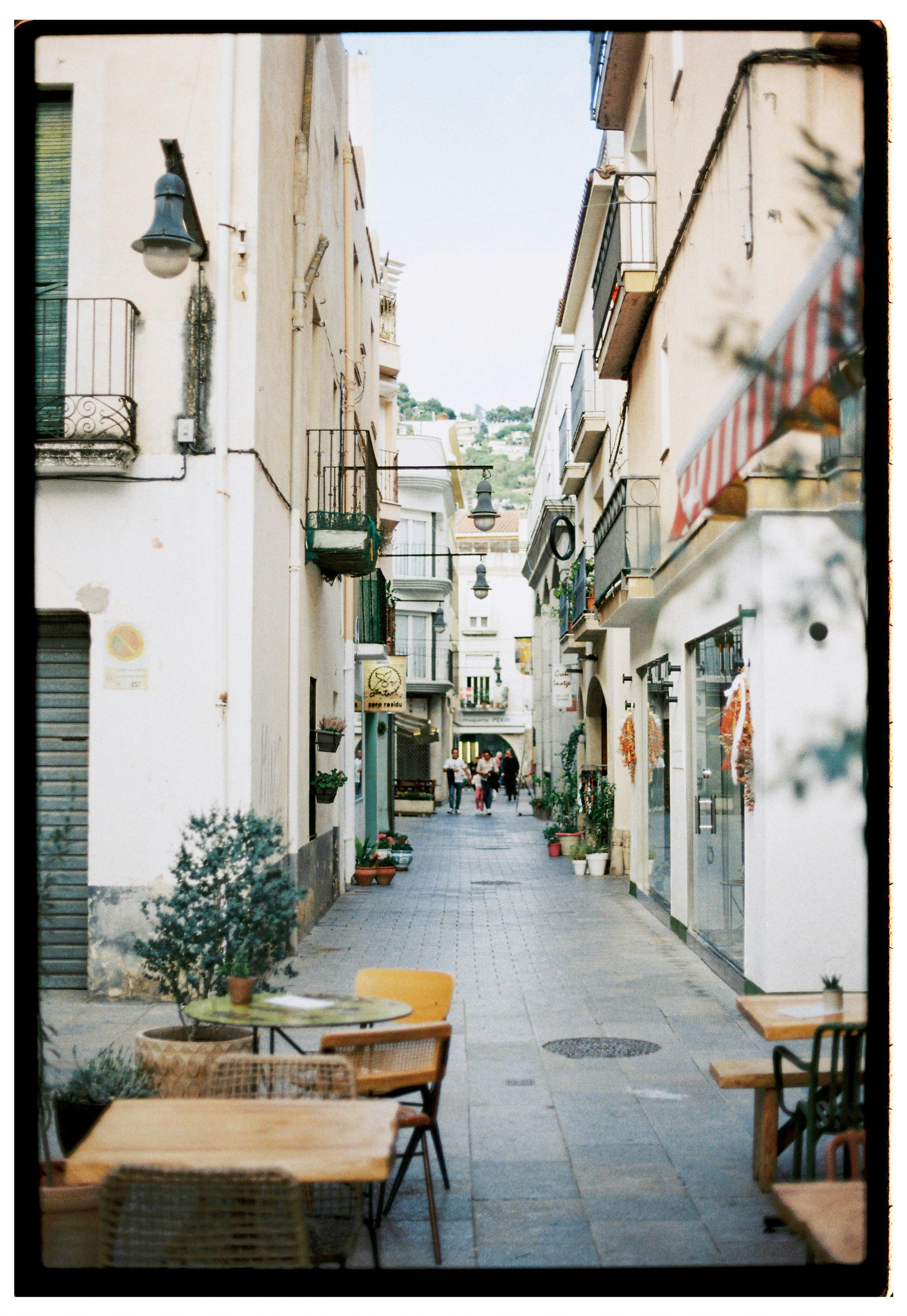Narrow Mediterranean street with outdoor cafe seating, capturing a relaxed, sunny atmosphere.