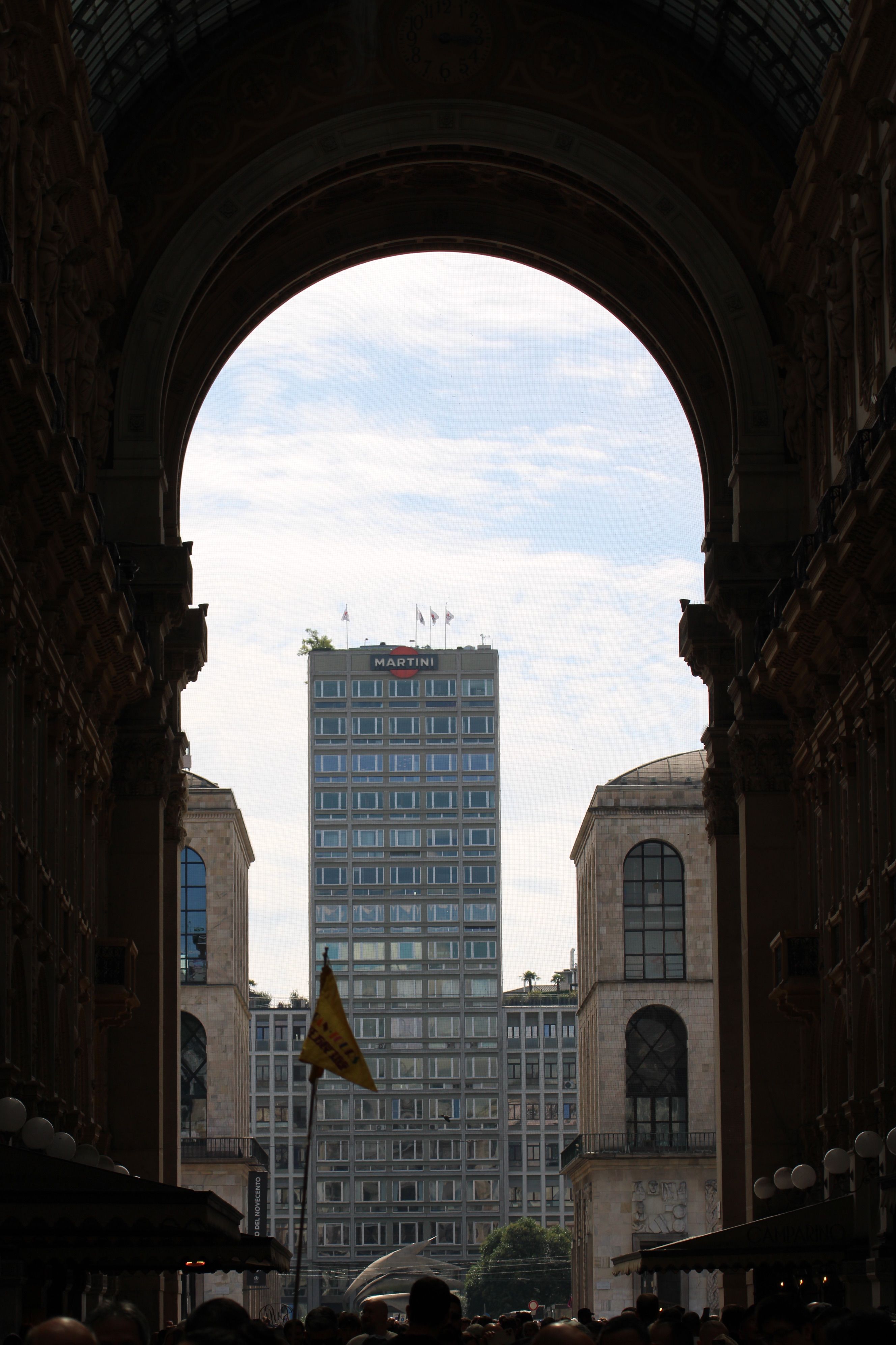 A tall skyscraper framed by the archway of Galleria Vittorio Emanuele II in Milan, Italy.