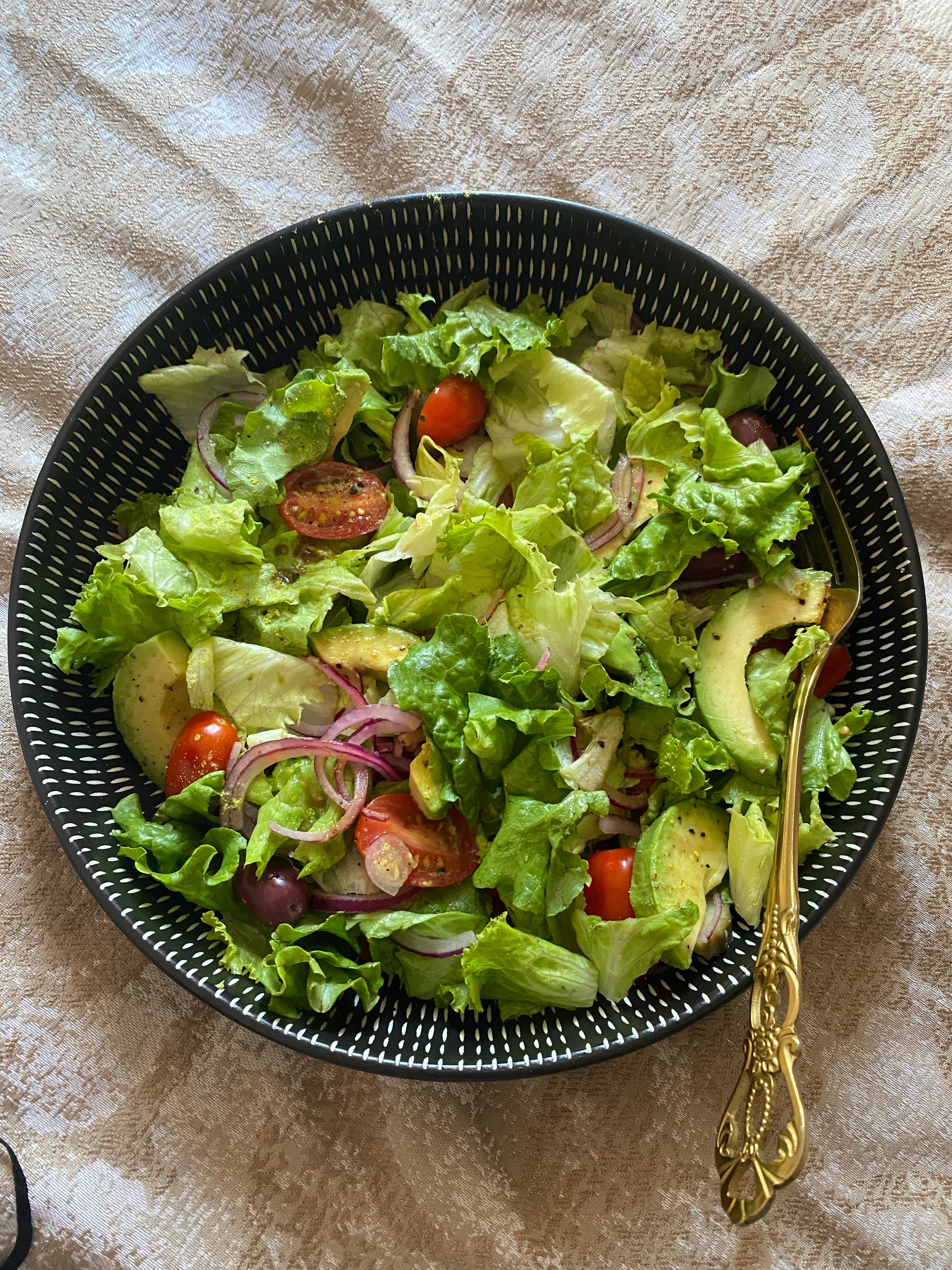 Healthy mixed veggie salad with avocado, lettuce, olives, and tomatoes in a black bowl.