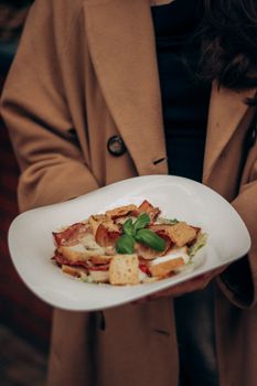 A stylish Caesar salad with basil leaves in an upscale dining environment, held by a person in a coat.