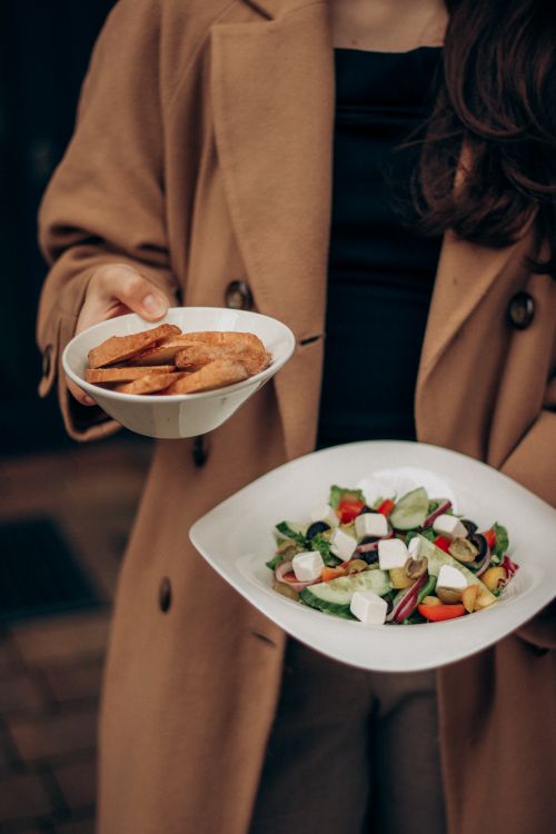 Free A woman in a beige coat holding a Greek salad and bread outside, showcasing fresh and healthy food. Stock Photo