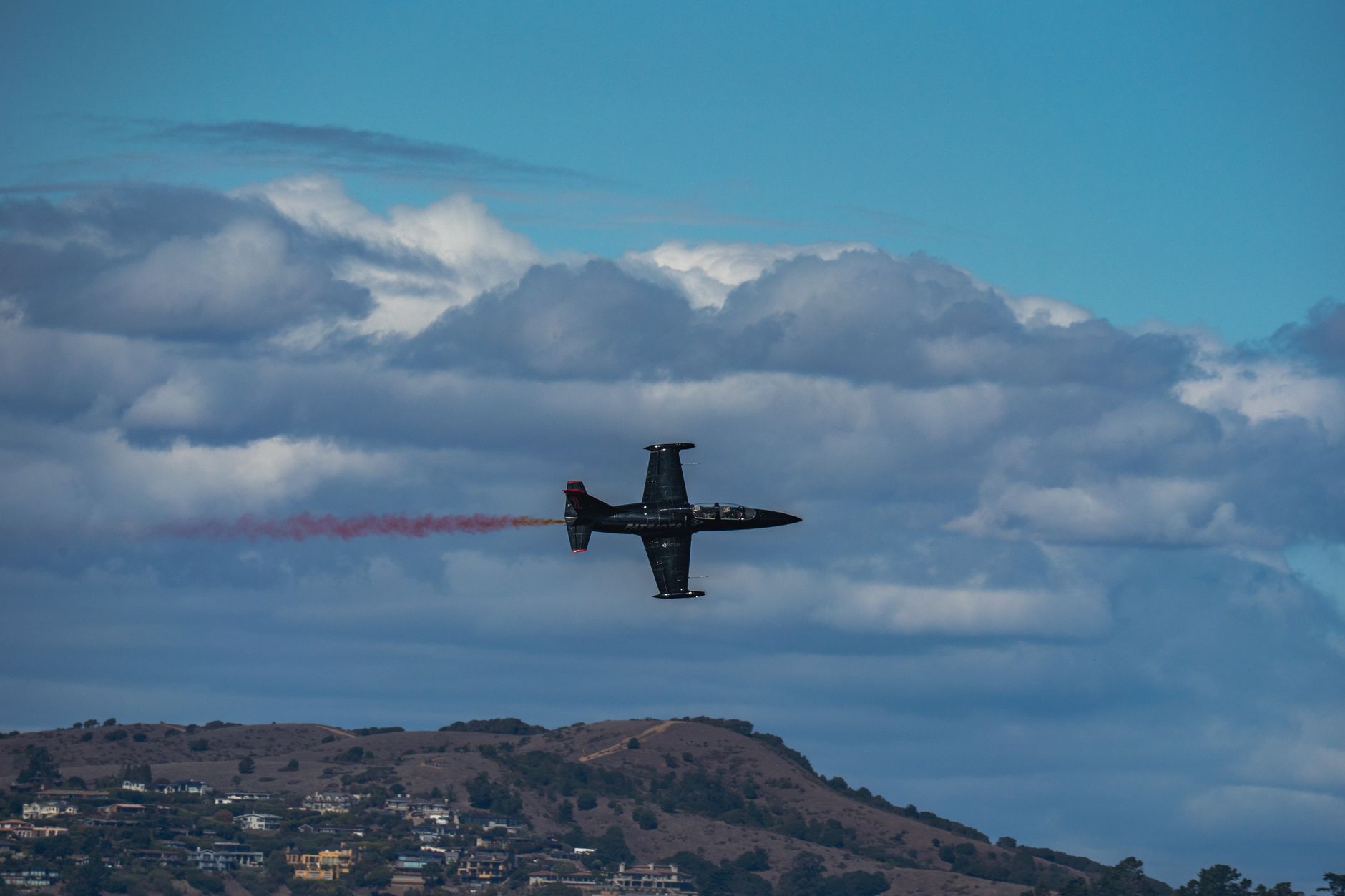 A fighter jet streaks across the sky leaving a trail, set against a backdrop of clouds and hills.