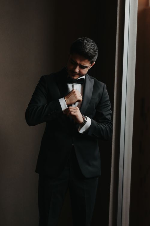 Free Sophisticated man in a tuxedo adjusting his cufflinks near a window, indoor setting. Stock Photo