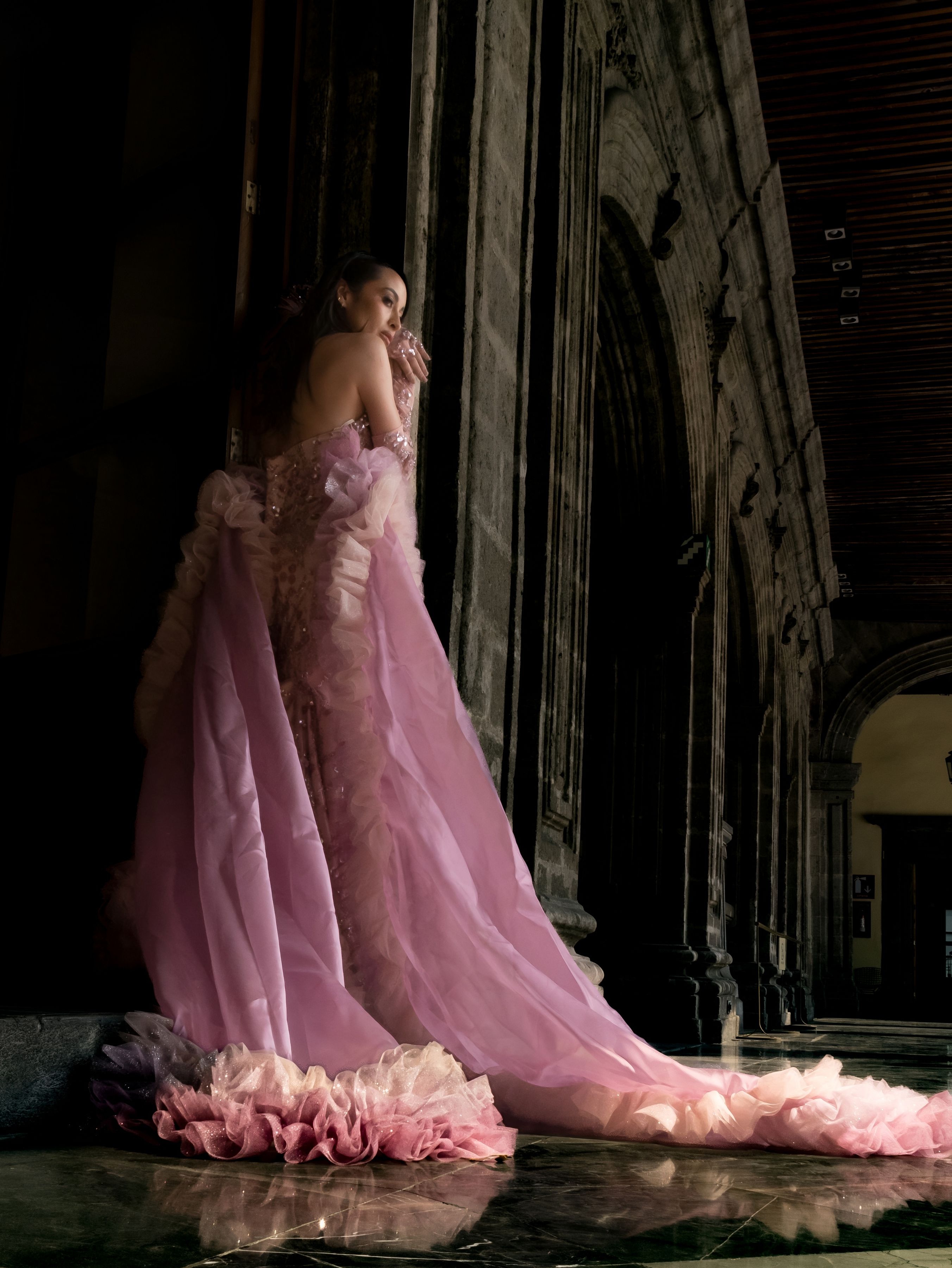 A woman in a flowing pink gown stands gracefully in a historic, dimly lit corridor.