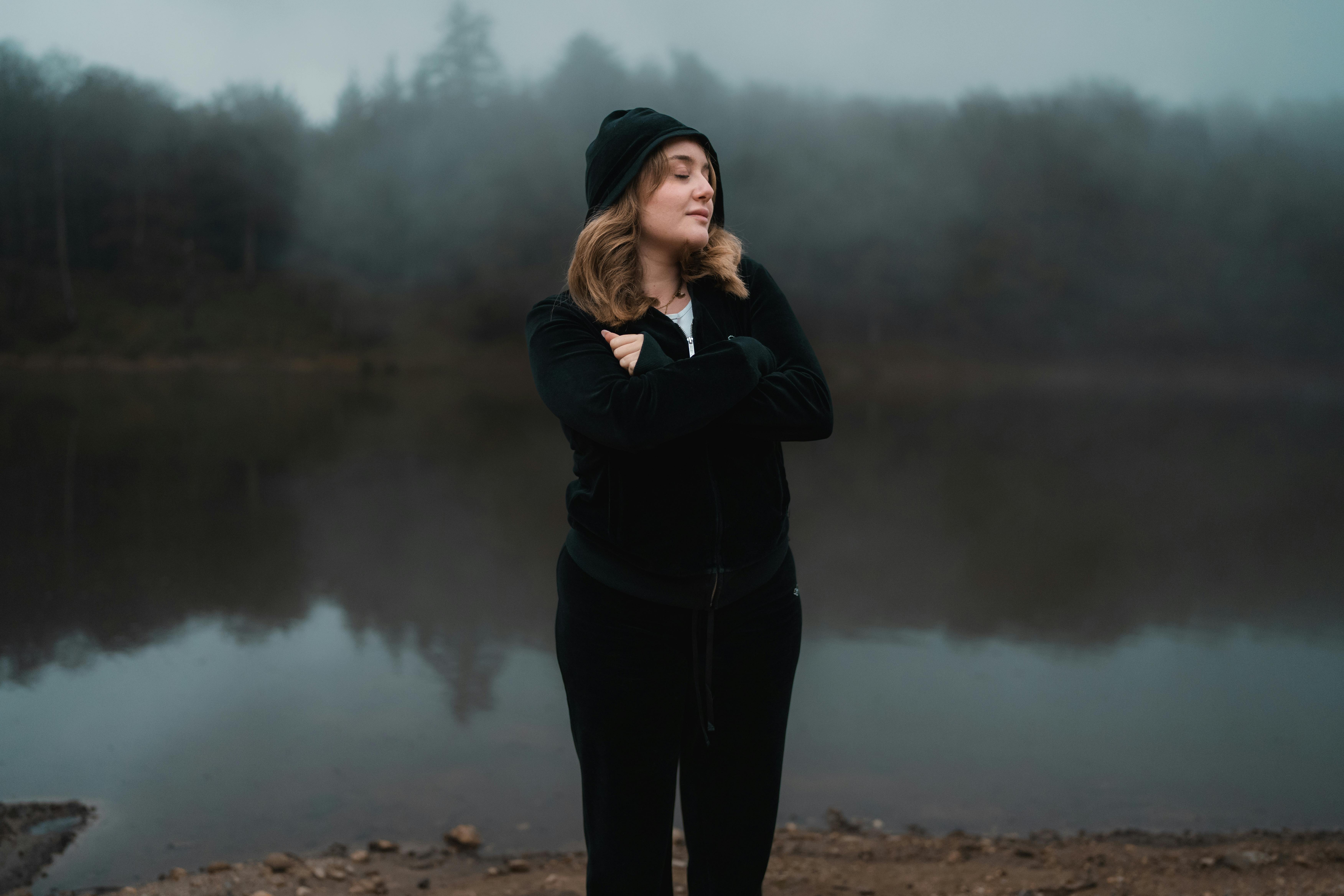 Une femme se tient contemplativement près d'un lac brumeux, entourée d'arbres brumeux.