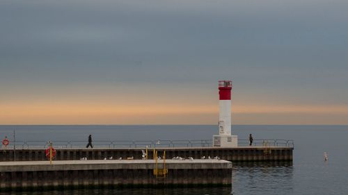 Free Lighthouse on a tranquil pier at sunset with a calm sea backdrop and silhouetted figures walking. Stock Photo
