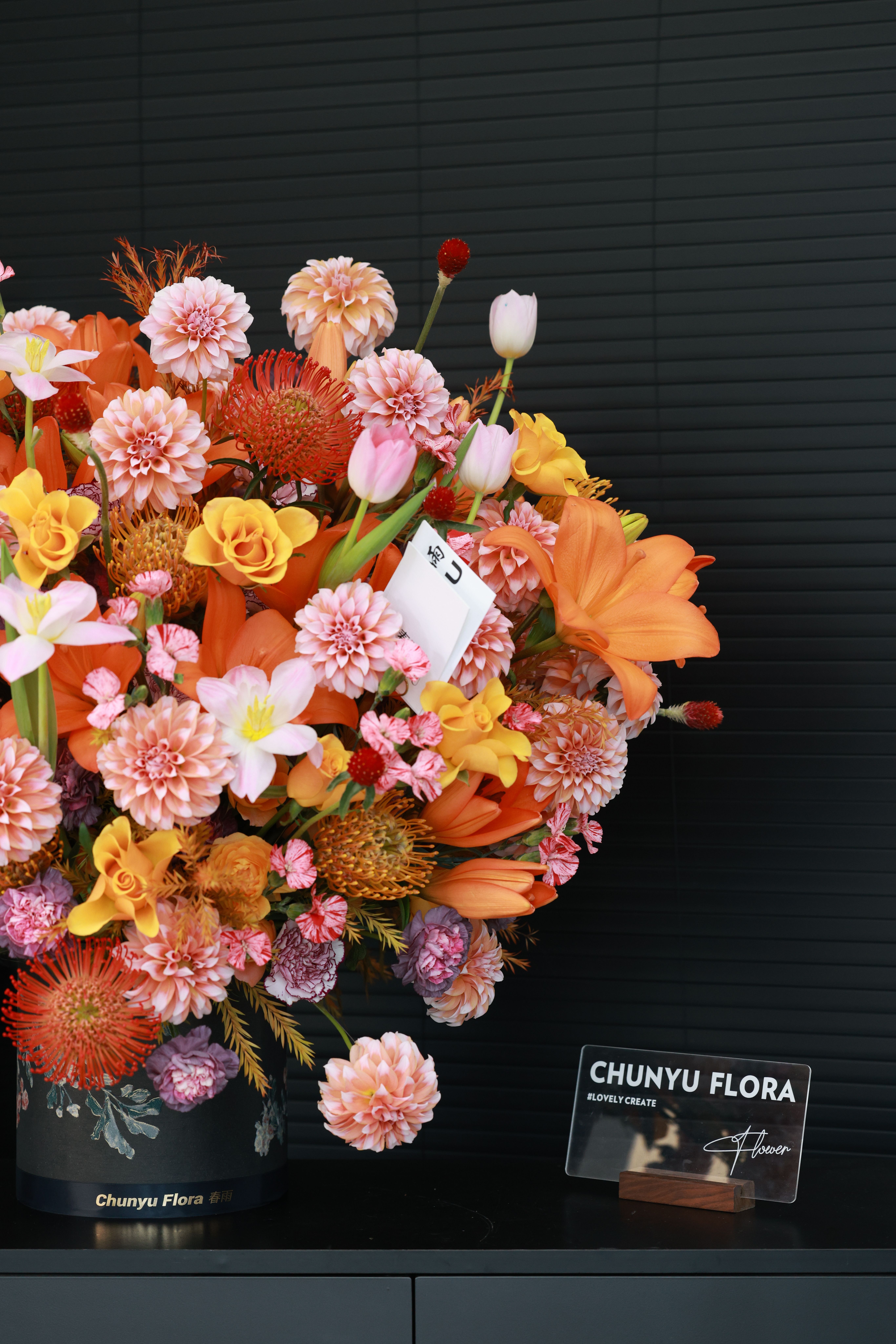 Colorful bouquet featuring dahlias, lilies, and roses in a decorative vase against a dark backdrop.