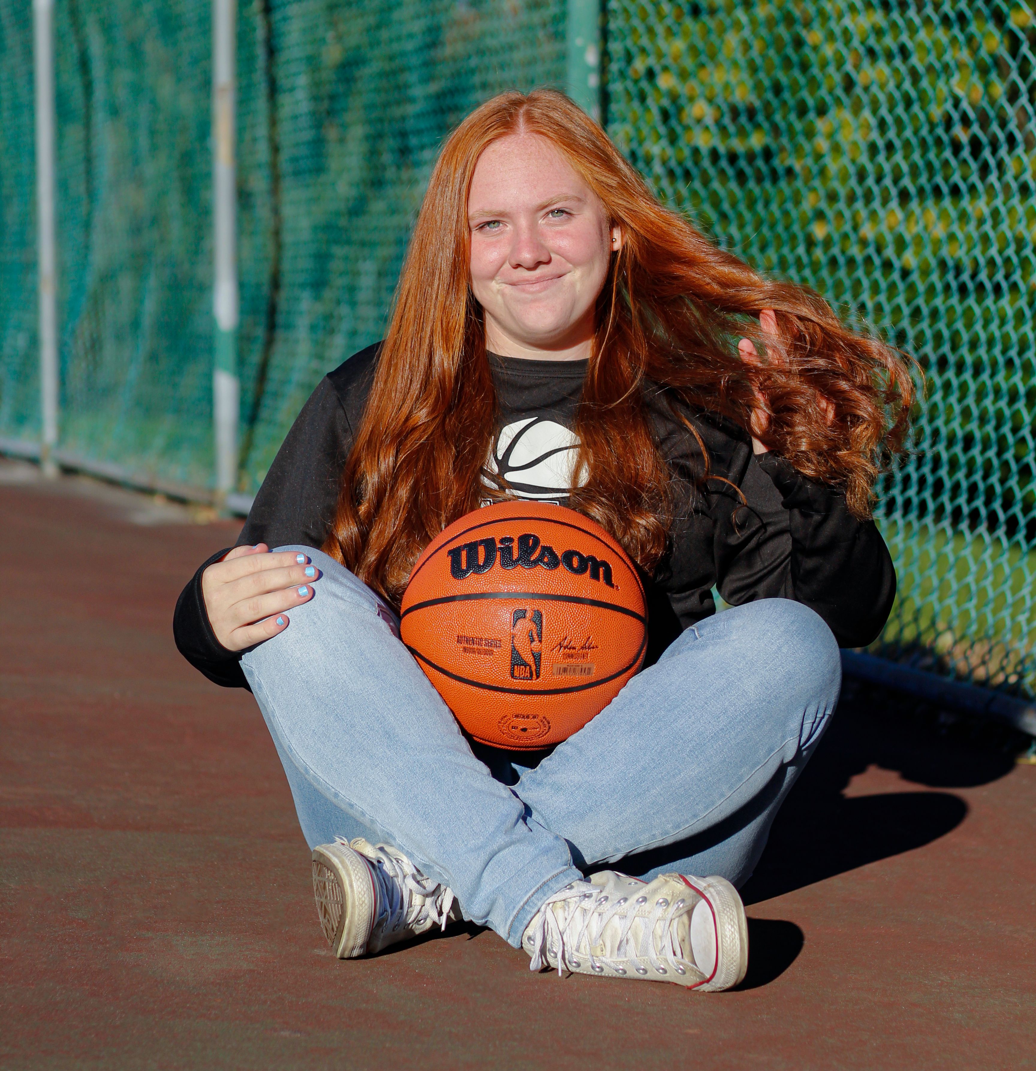 Smiling teen girl with red hair holding a Wilson basketball on an outdoor court.