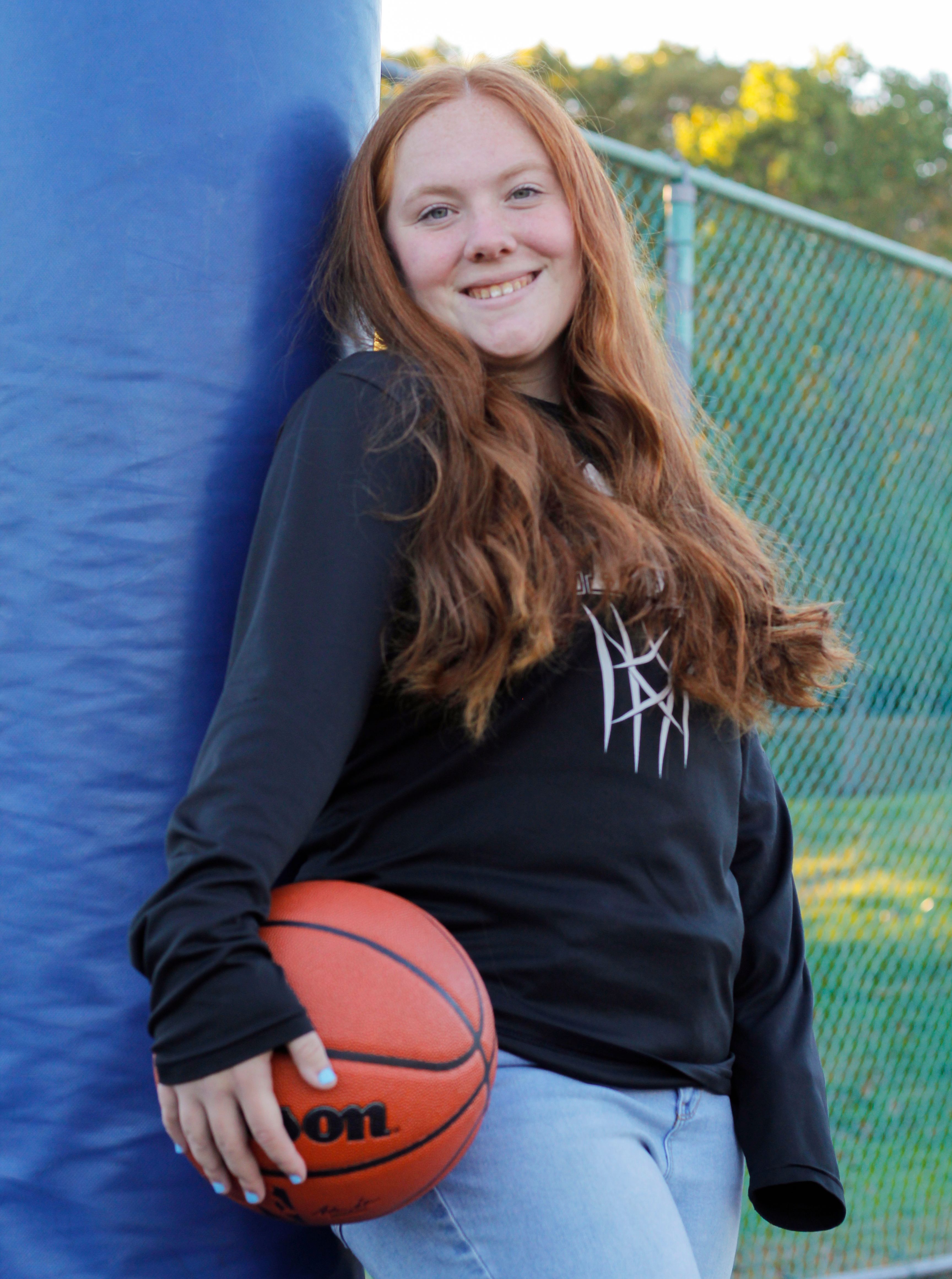 Smiling teenage girl leaning against pole with basketball outdoors. Casual and sporty vibe.