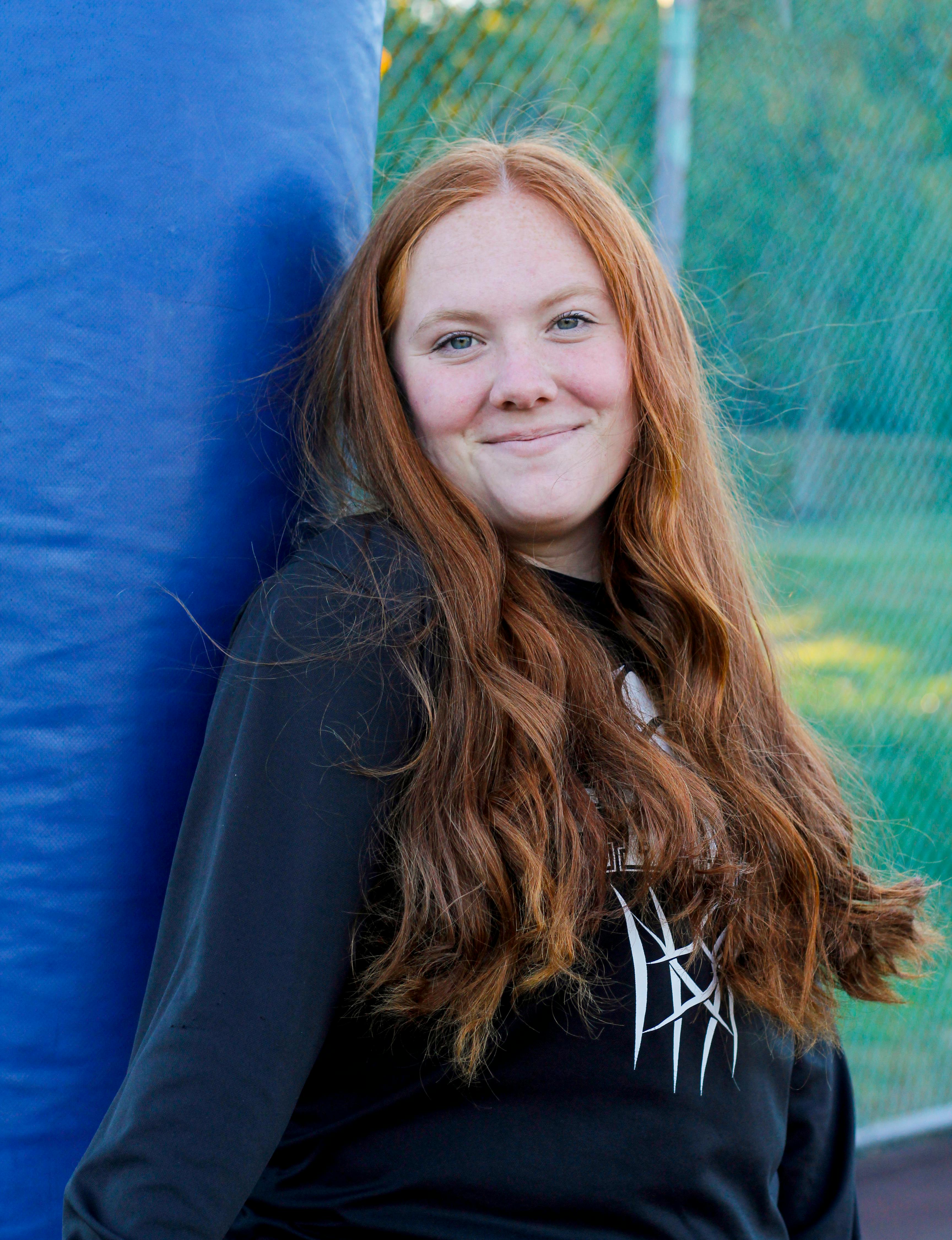 Young woman smiling, leaning on blue post outdoors, with long red hair and casual clothing.