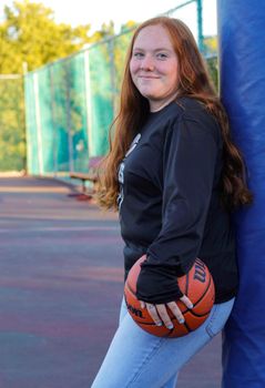 Smiling teenage girl leaning against a pole holding a basketball on an outdoor court.