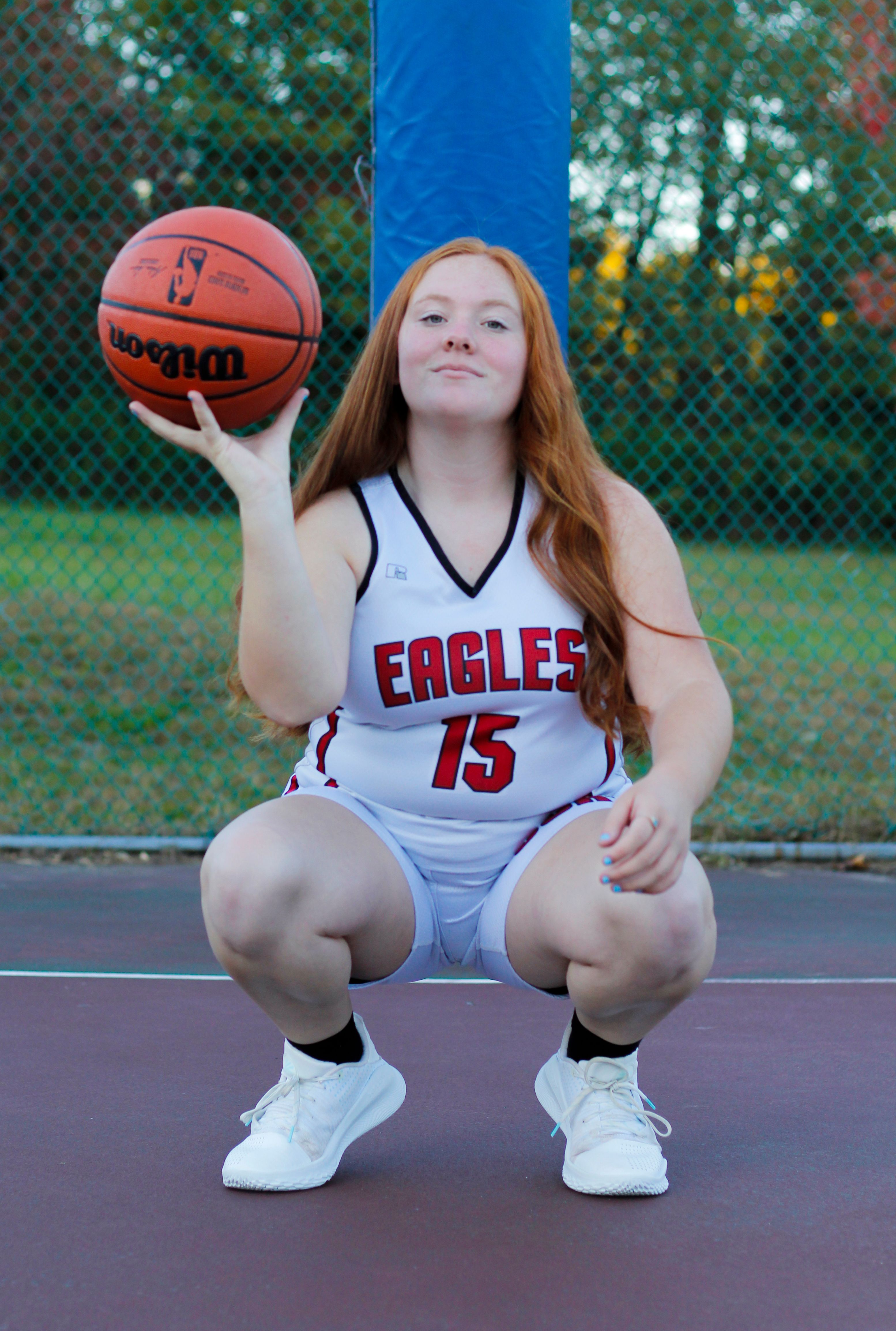 Teen basketball player posing on an outdoor court with a basketball, wearing a team jersey.
