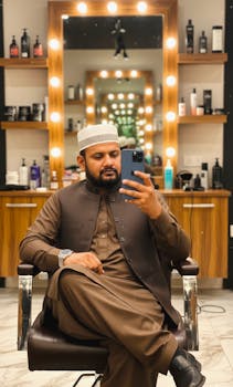 A man wearing traditional attire takes a selfie in a stylish barbershop with illuminated mirrors.
