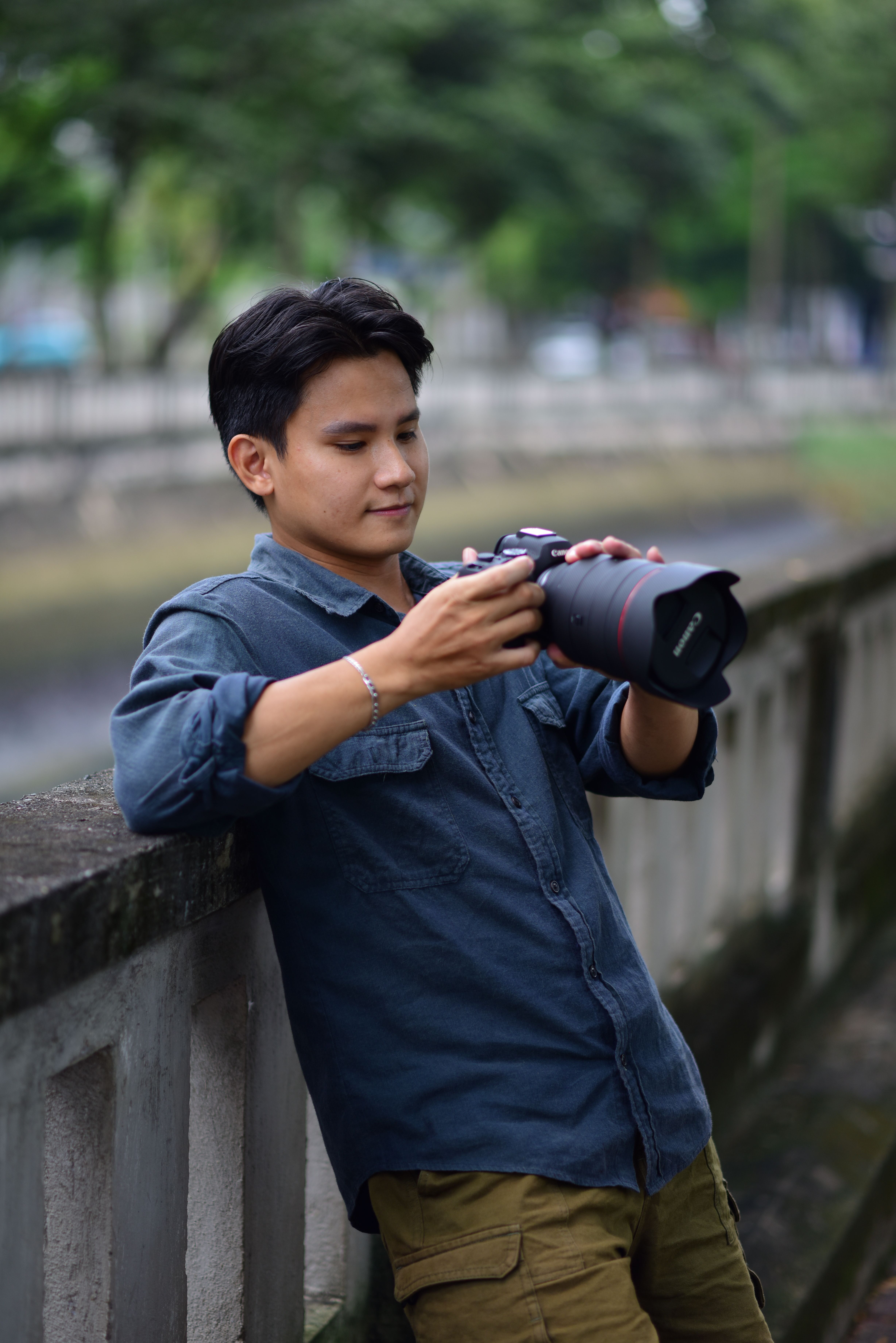 A young adult male photographer leaning on a railing checks his camera settings outdoors.