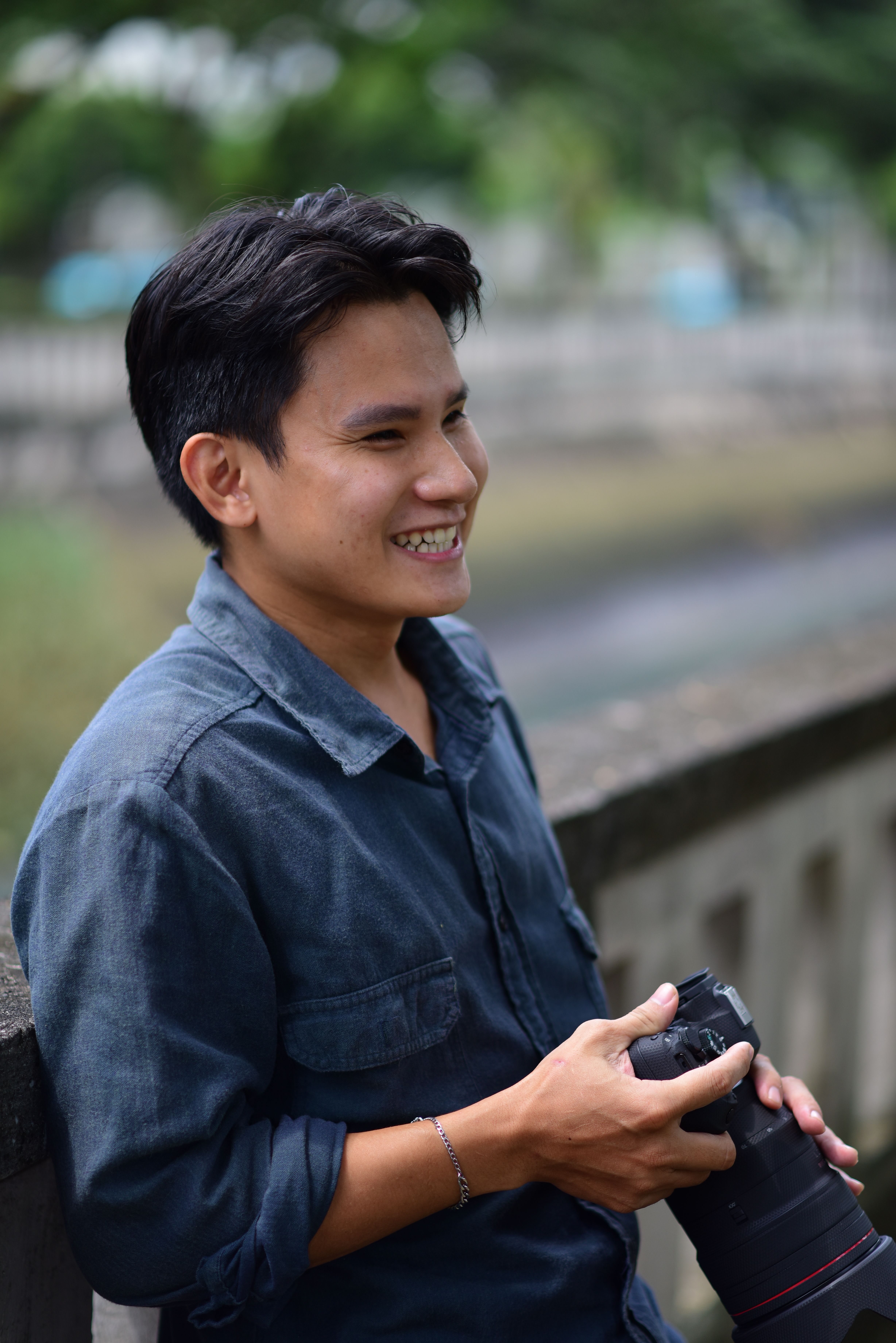 Young man holding a camera and smiling while leaning on a railing outdoors.