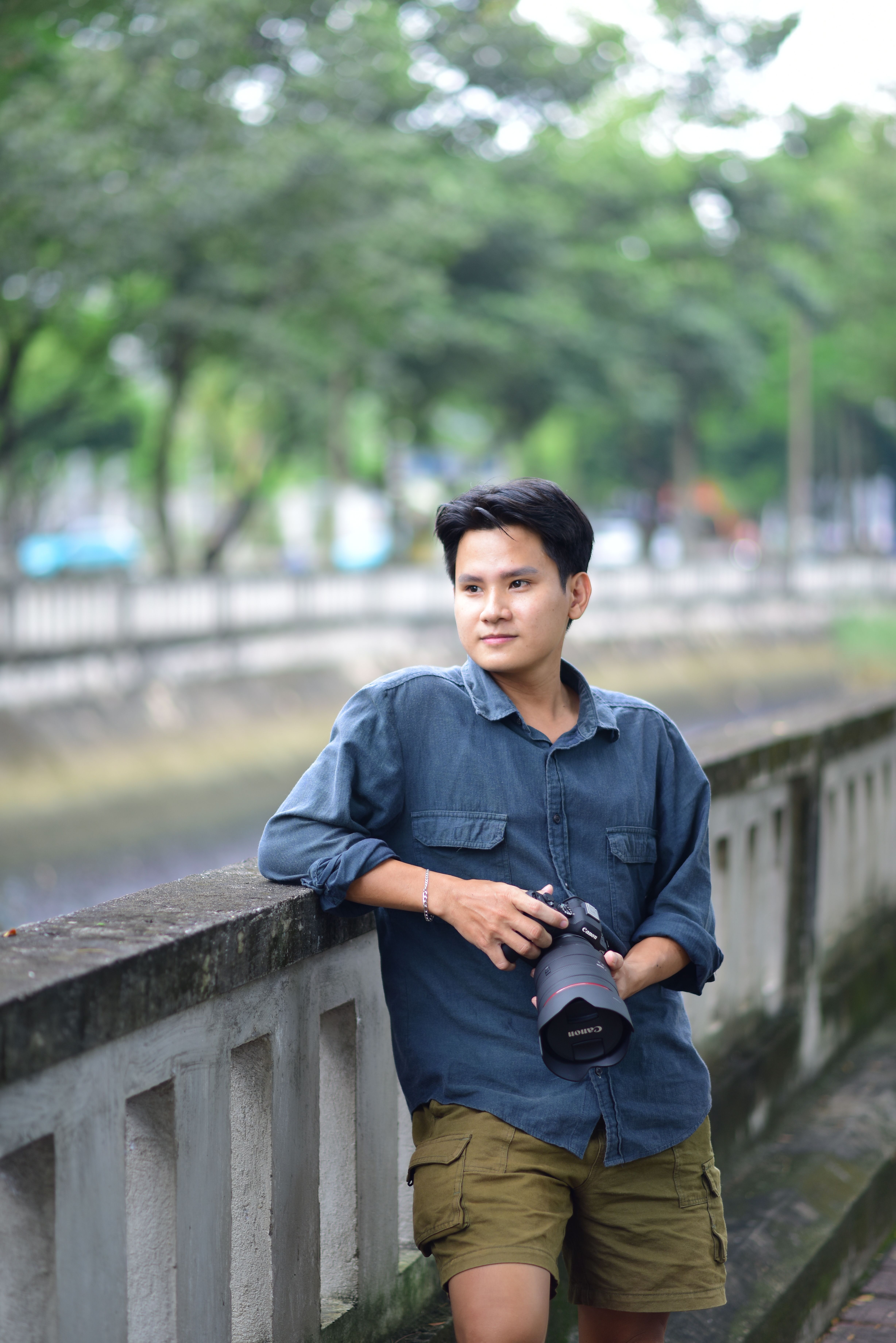 Casual portrait of a man leaning on a bridge holding a camera by a canal.