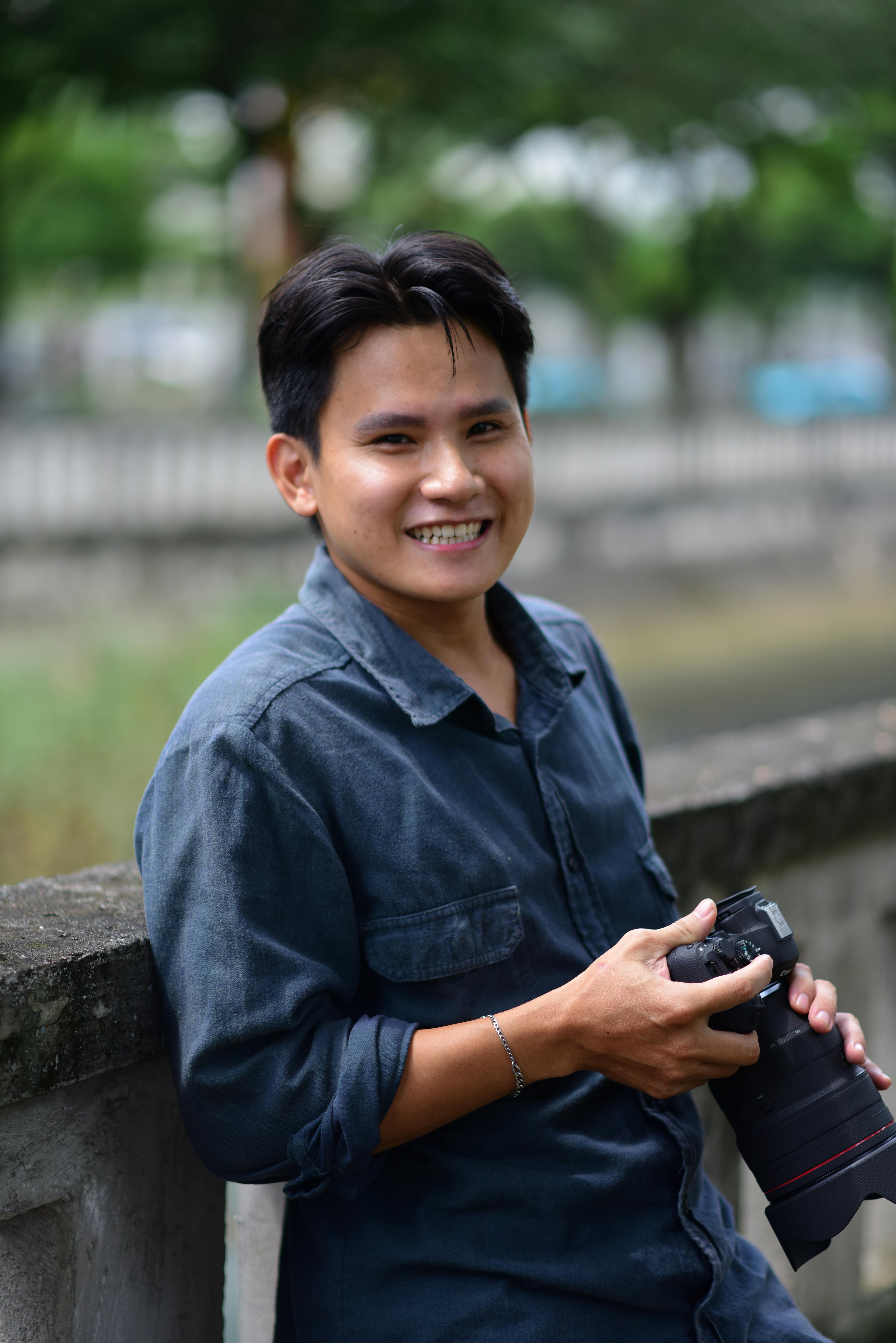 Miễn phí Người đàn ông mỉm cười cầm máy ảnh, tạo dáng ngoài trời. Bối cảnh tự nhiên. Ảnh lưu trữ