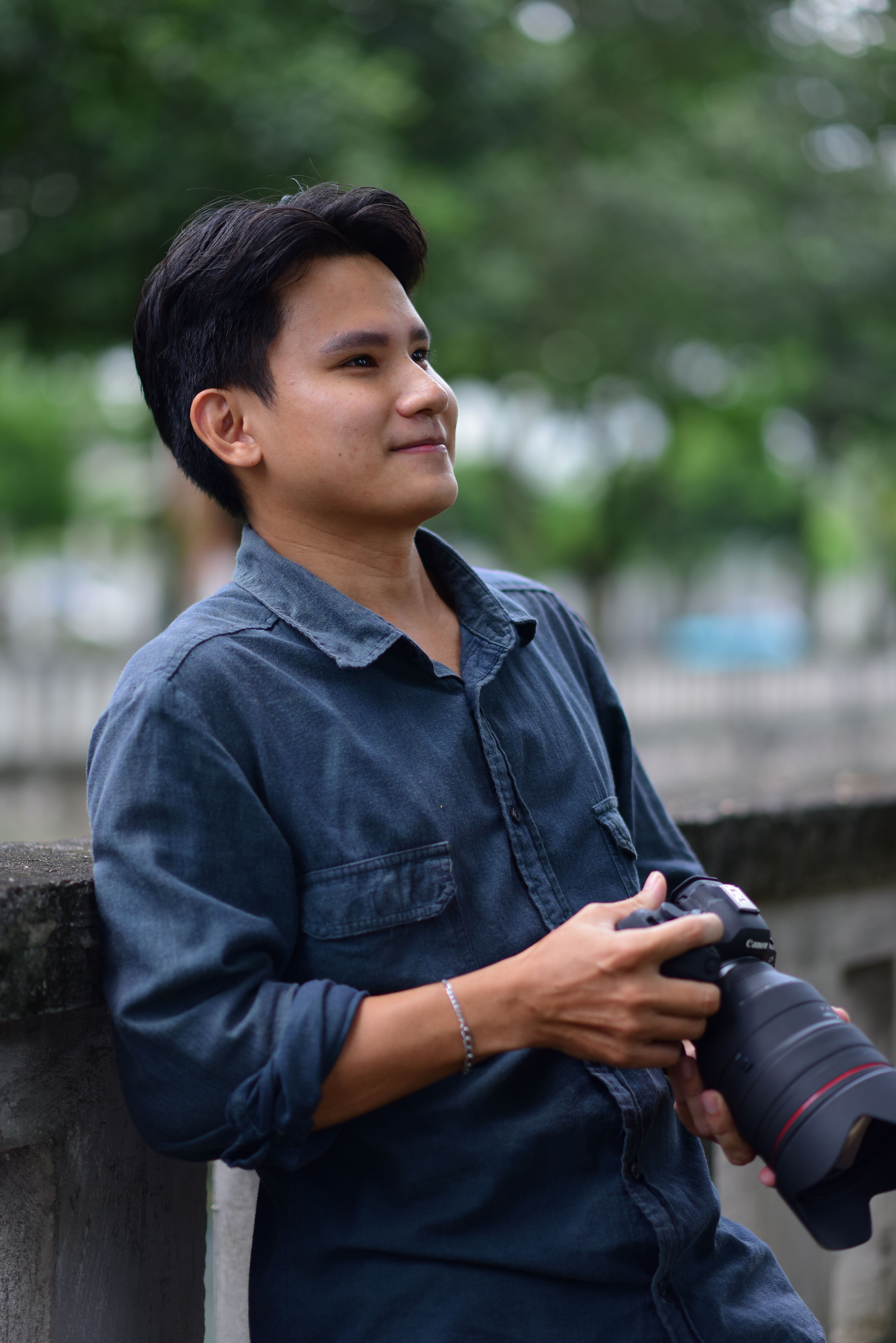 Young male photographer holding a camera, posing casually outdoors in a park setting.