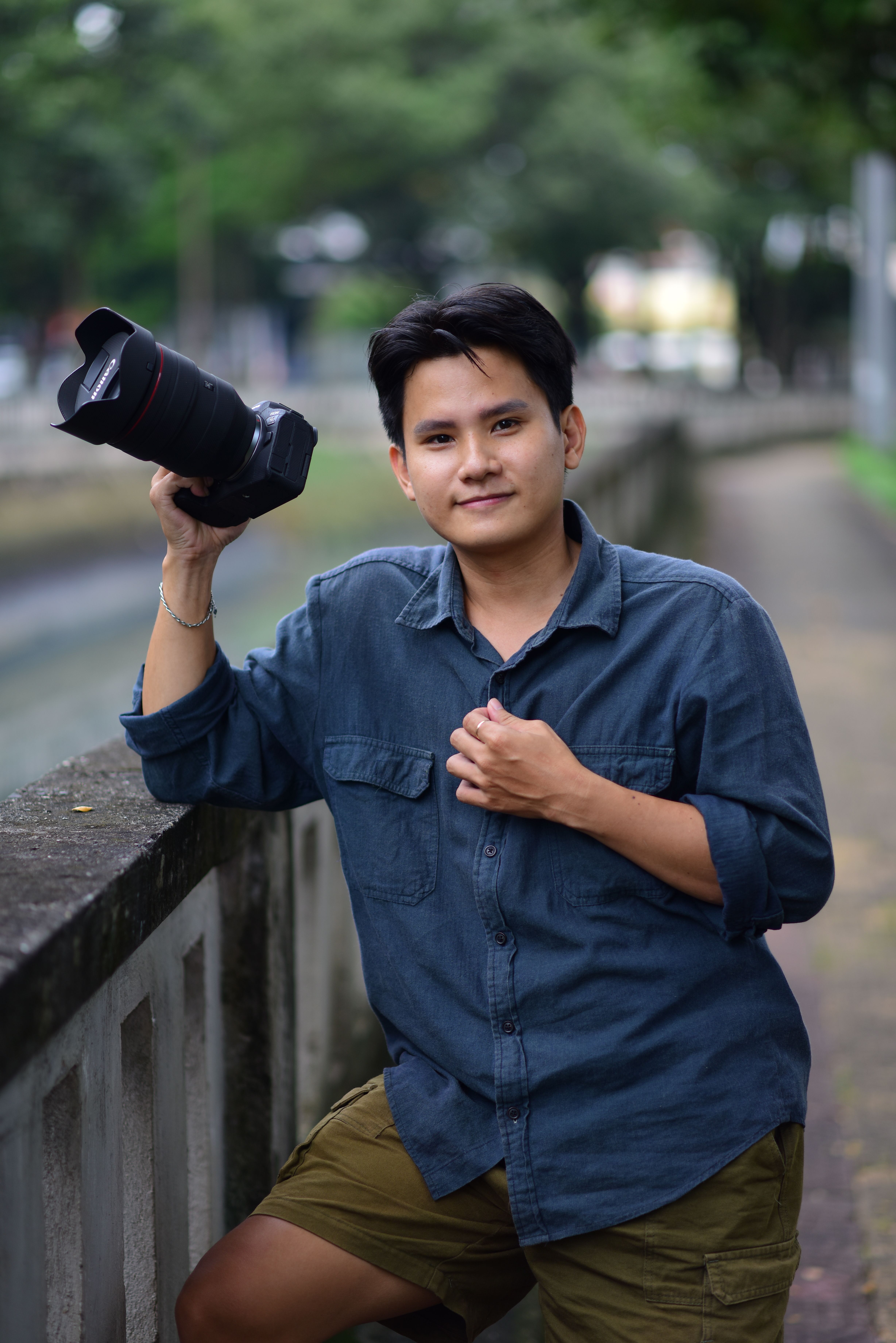 Young male photographer poses with a camera outdoors, showcasing casual style and expertise.