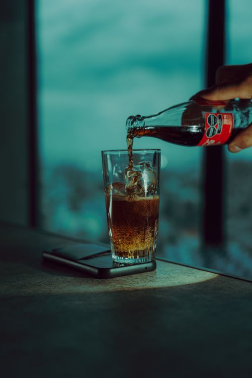 Free A person pours cola into a glass placed on a table with a smartphone nearby in a modern setting. Stock Photo