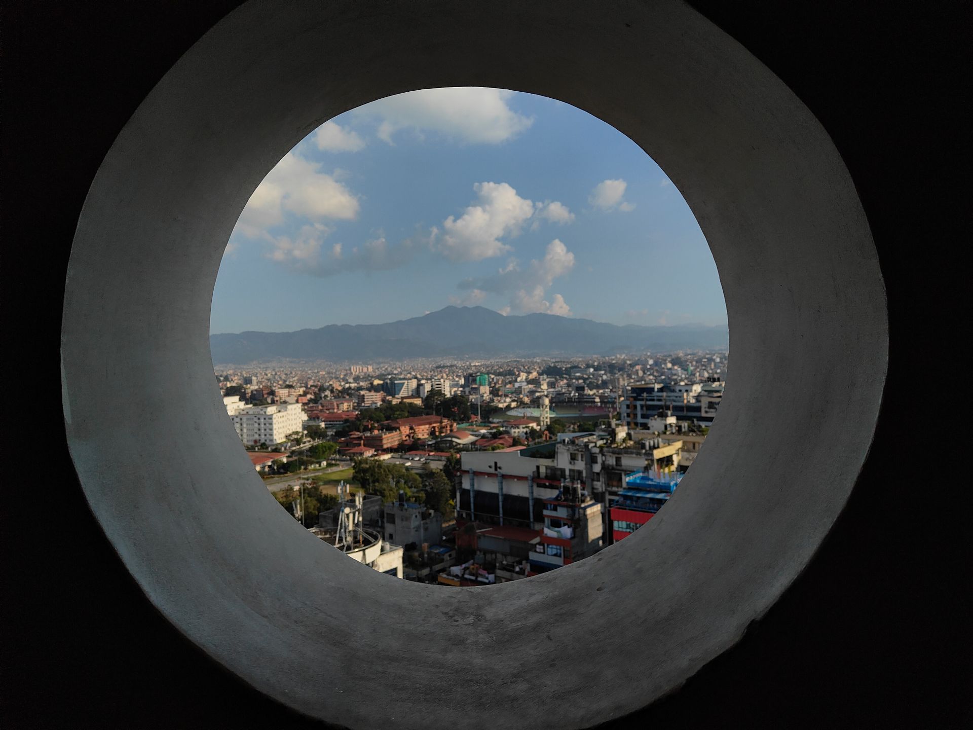 Unique cityscape view framed by circular window, mountains in the background.