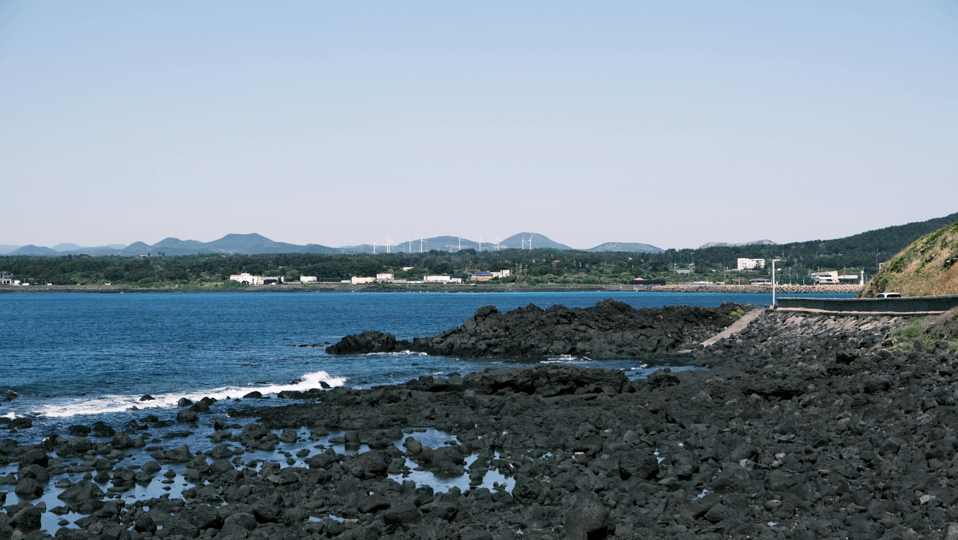 Explore the rocky coastline and distant wind turbines of Jeju Island, offering a serene scenic view.