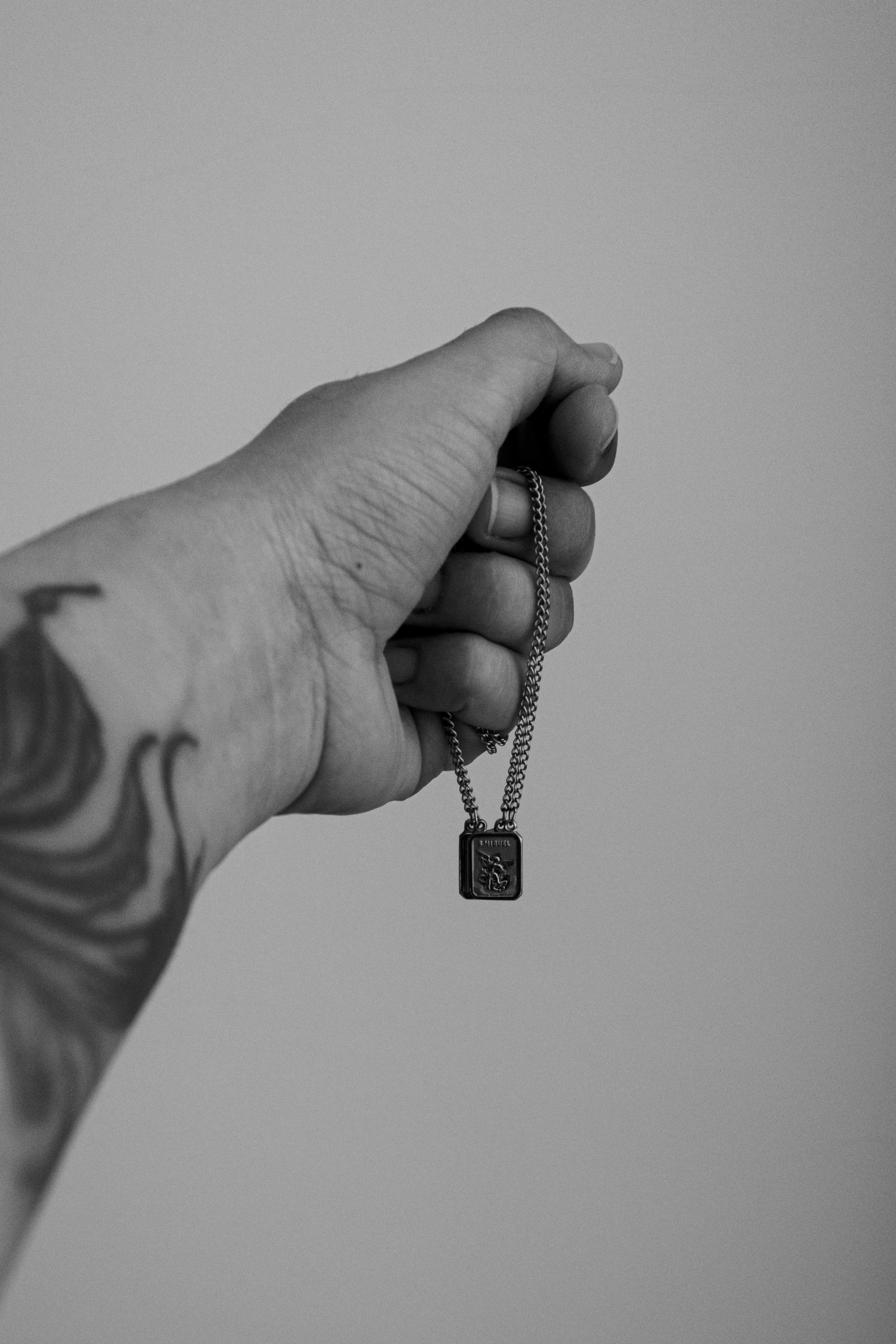 Gratis En tatovert hånd som holder et anhengskjede mot en vanlig bakgrunn, fanget i svart-hvitt. Arkivbilde