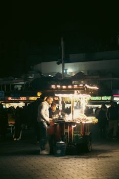 Street vendor selling illuminated snacks at night market surrounded by crowd.