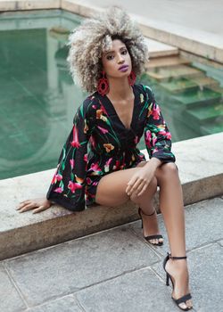 Fashionable Afro-Brazilian woman seated by a pool in a vibrant floral dress, exuding elegance.