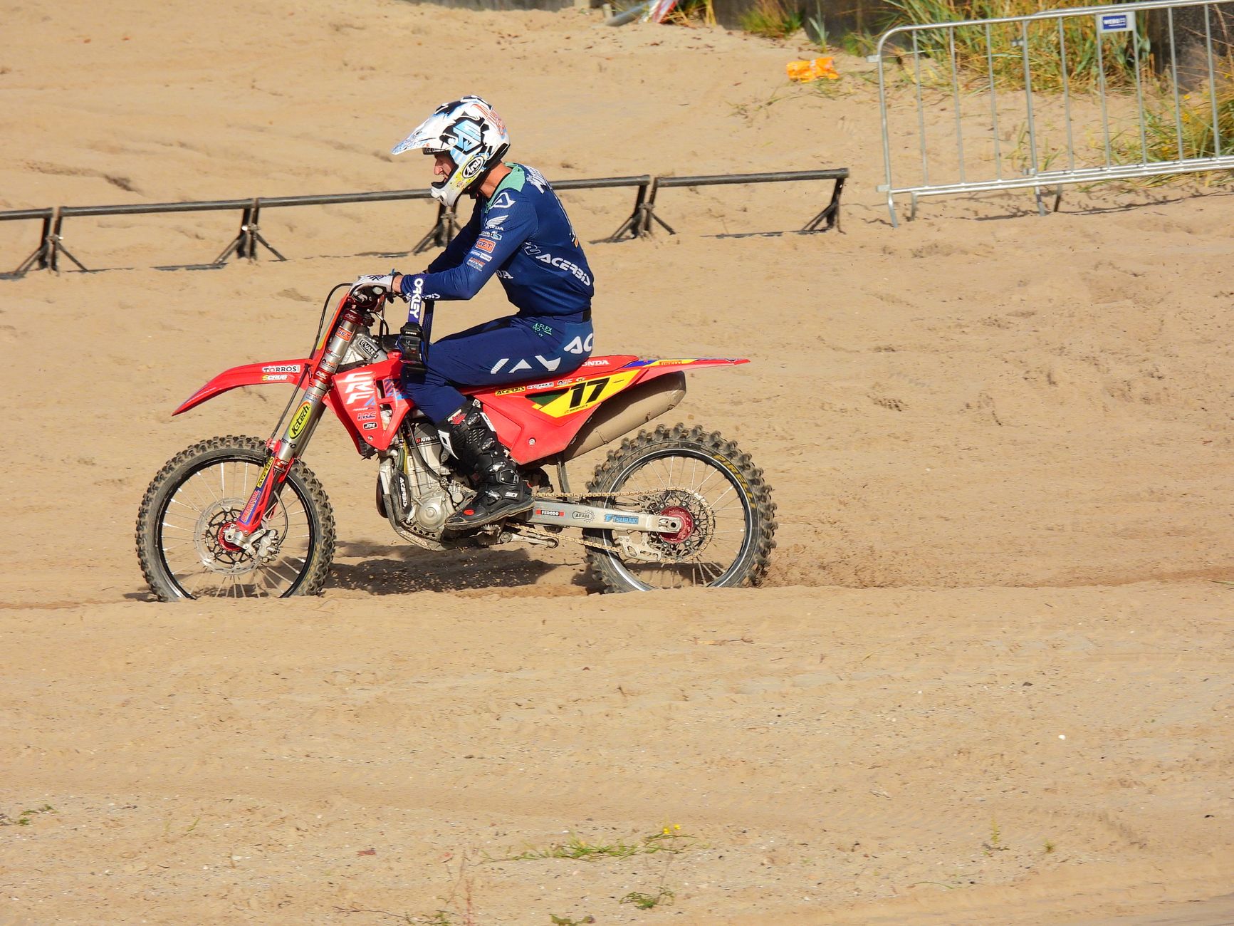 Motocross rider in full gear riding on sandy terrain during a daylight race event.