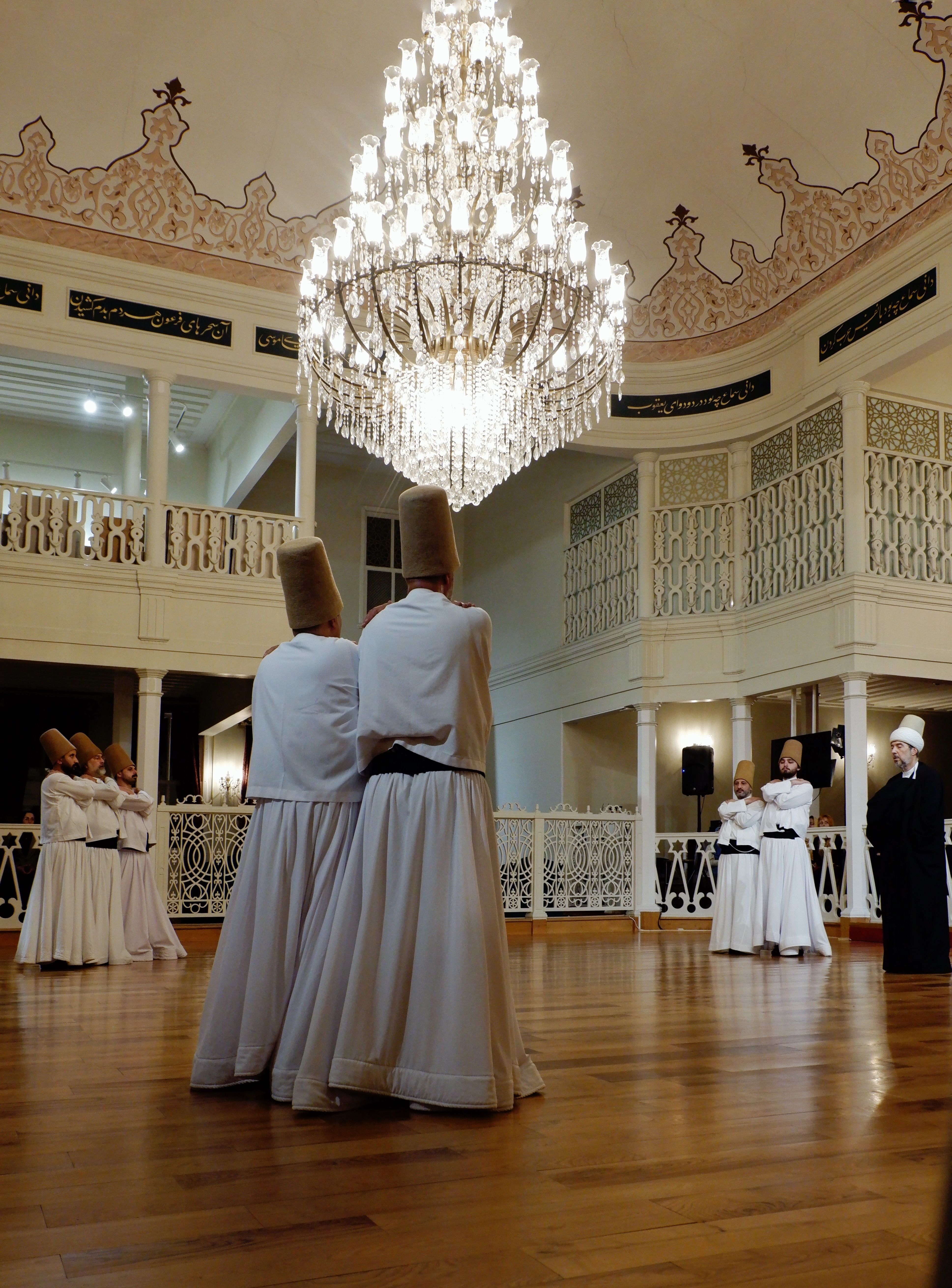 Whirling Dervishes performing a spiritual dance under a grand chandelier in an ornate hall.