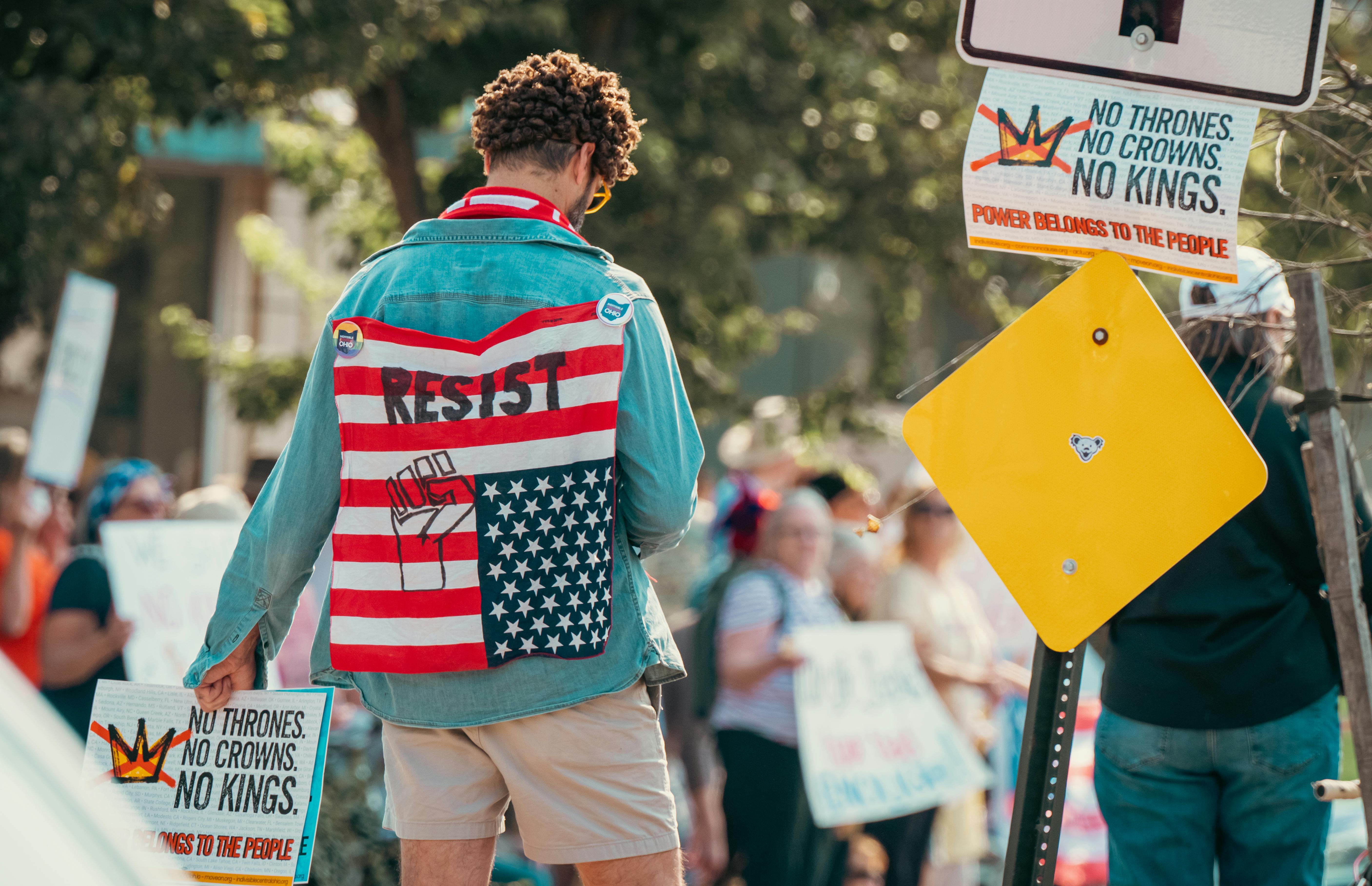 Person wearing flag-themed denim jacket at a protest holding signs promoting resistance and anti-est