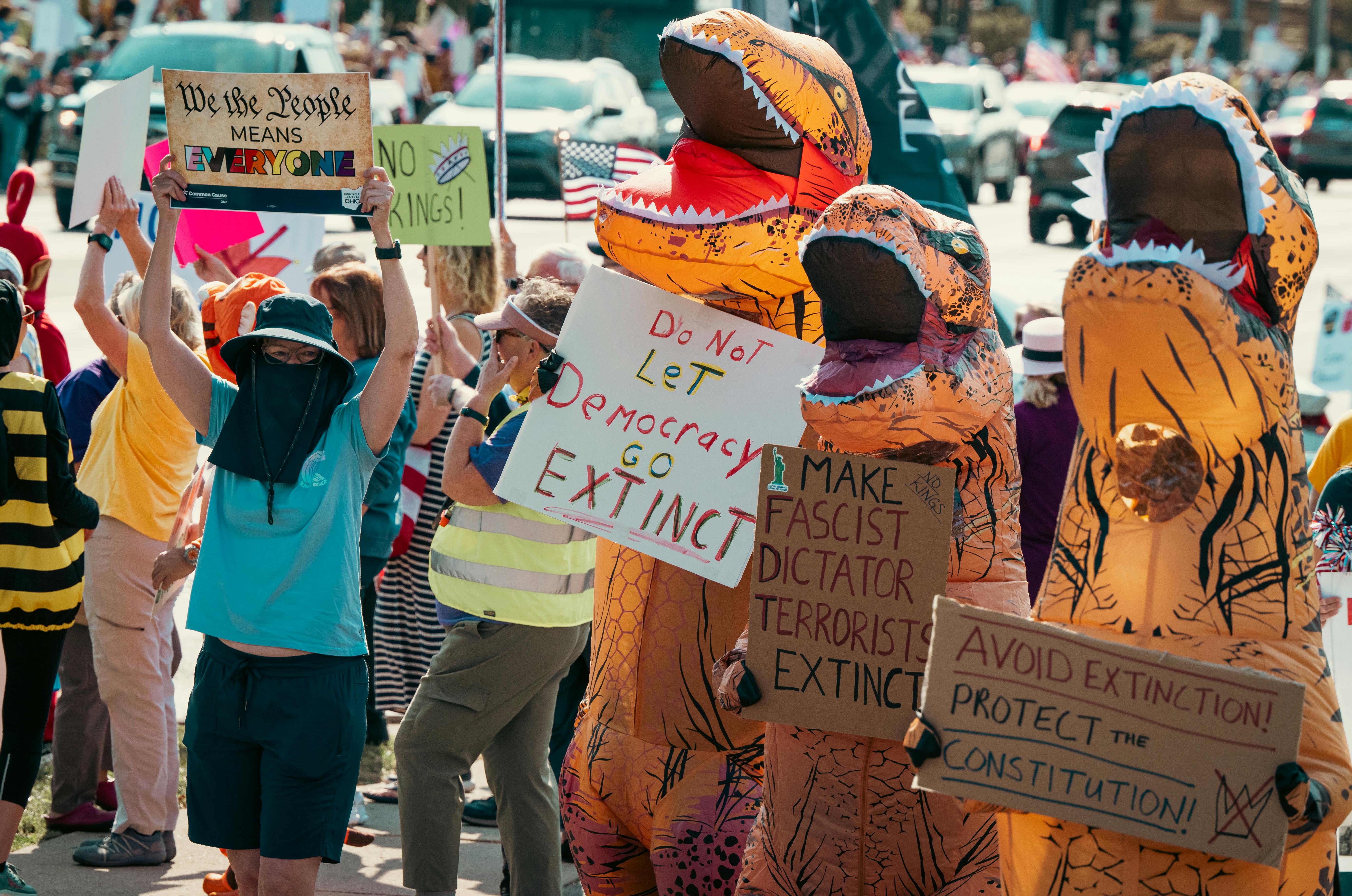 Protesters dressed in dinosaur costumes hold signs advocating for democracy during a vibrant outdoor rally.