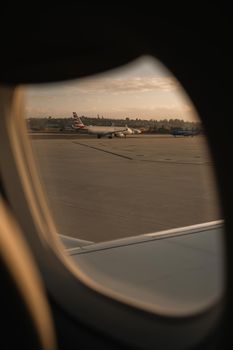 View from airplane window showing airport runway and planes during sunset.